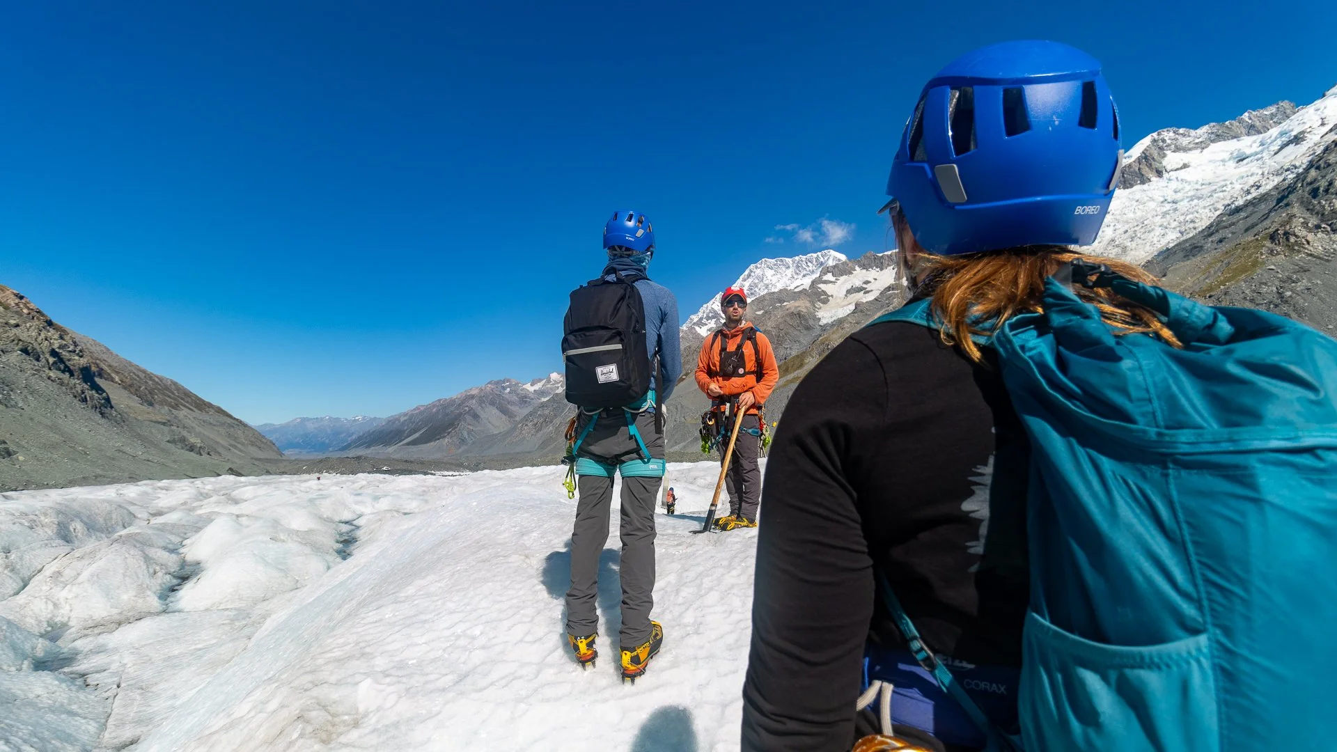 Group of four people standing on snow with mountain background, wearing helmets and outdoor hiking gear in a mountainous landscape.