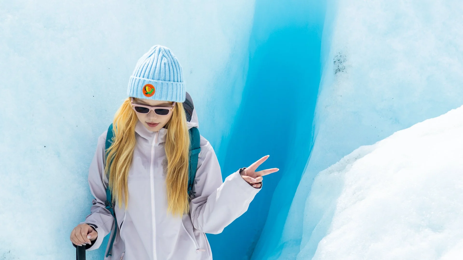 A woman with long blonde hair in a white jacket, wearing sunglasses, a blue beanie with a colorful patch, and a blue backpack, standing in an ice cave. She is making a peace sign with her right hand, surrounded by blue ice walls and snow.