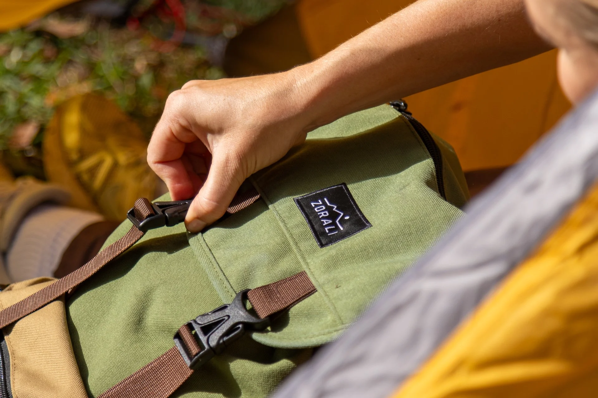 Close-up of a person's hand adjusting black strap on a green backpack with a logo that says 'ZOMAL' in white letters, outdoors with grass and leaves in the background.
