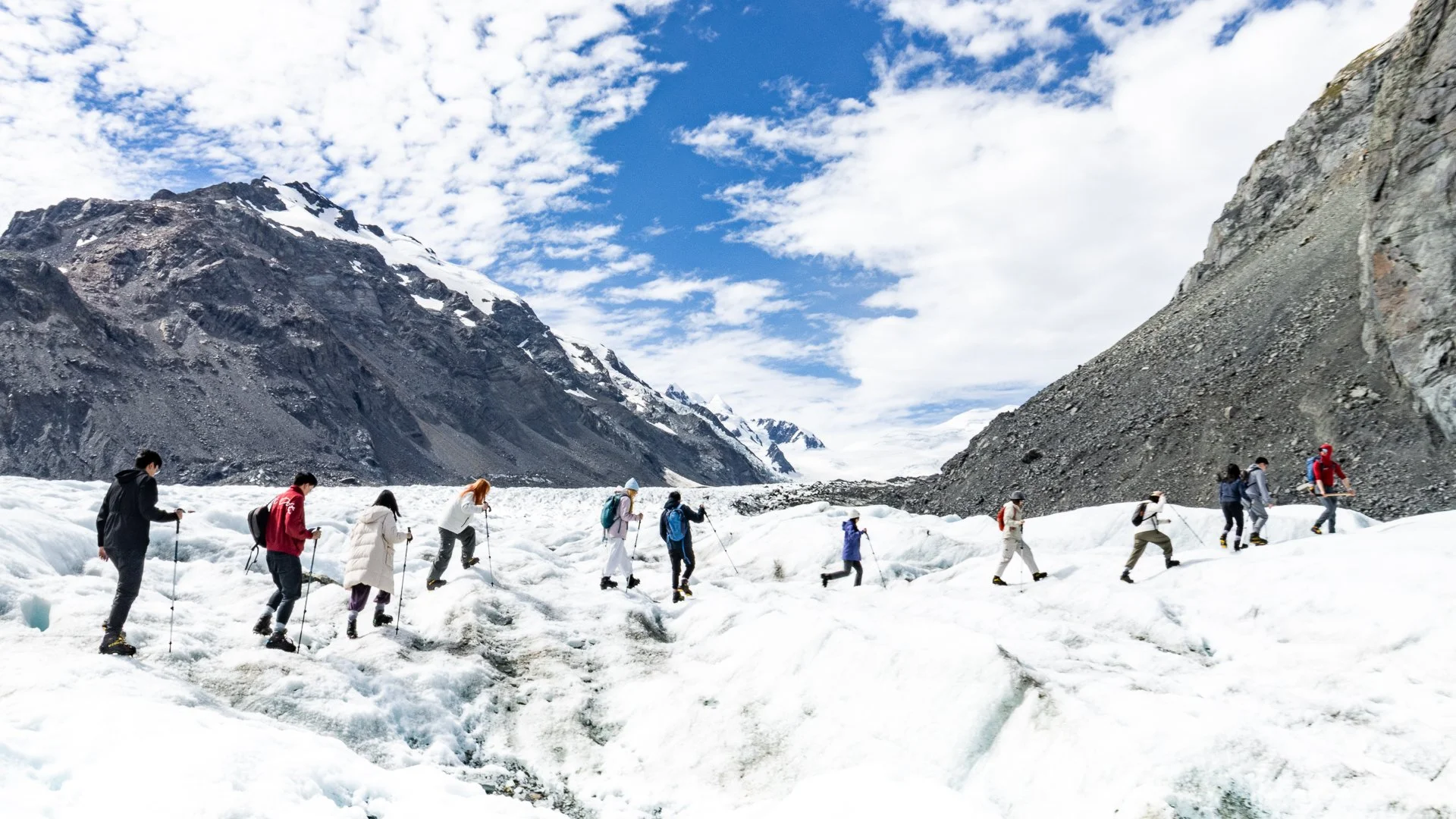 A group of hikers trekking across a snowy glacier in a mountainous landscape with steep rocky slopes and a partly cloudy sky.