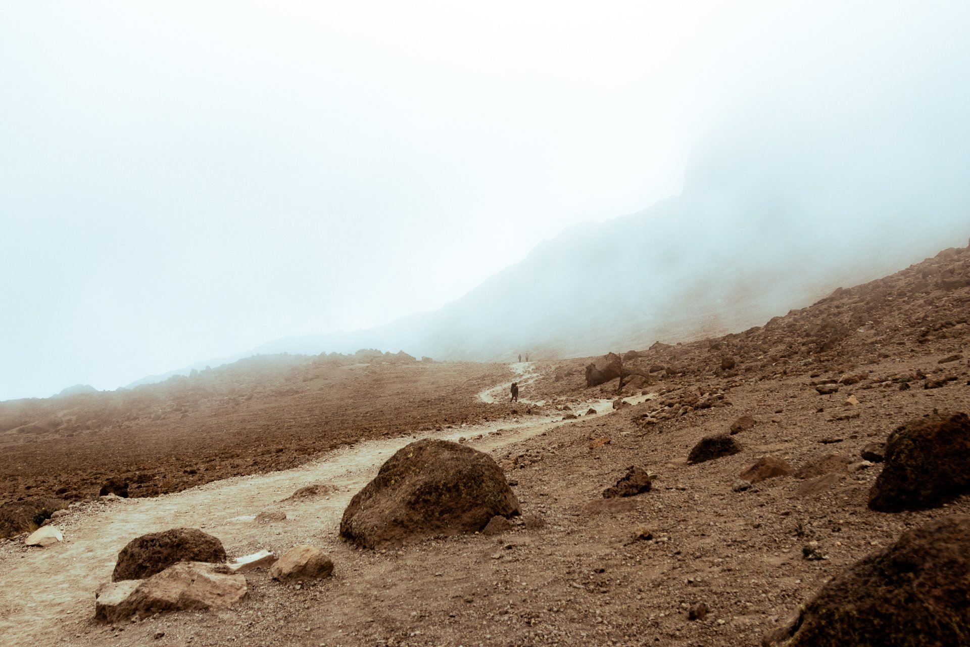 Sandy, rocky terrain with a mountain in the background, covered in fog, and a few scattered hikers walking along a trail.