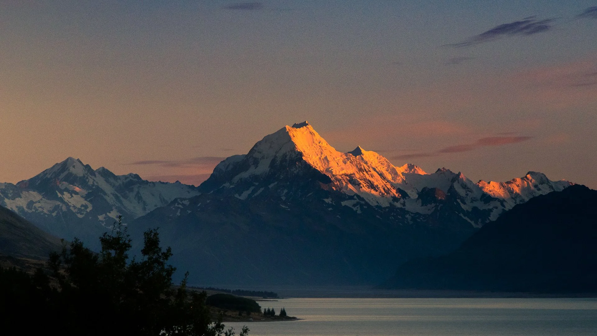 Snow-capped mountain peaks at sunset with the tallest peak illuminated in orange, calm water in the foreground, and a cloudy sky.