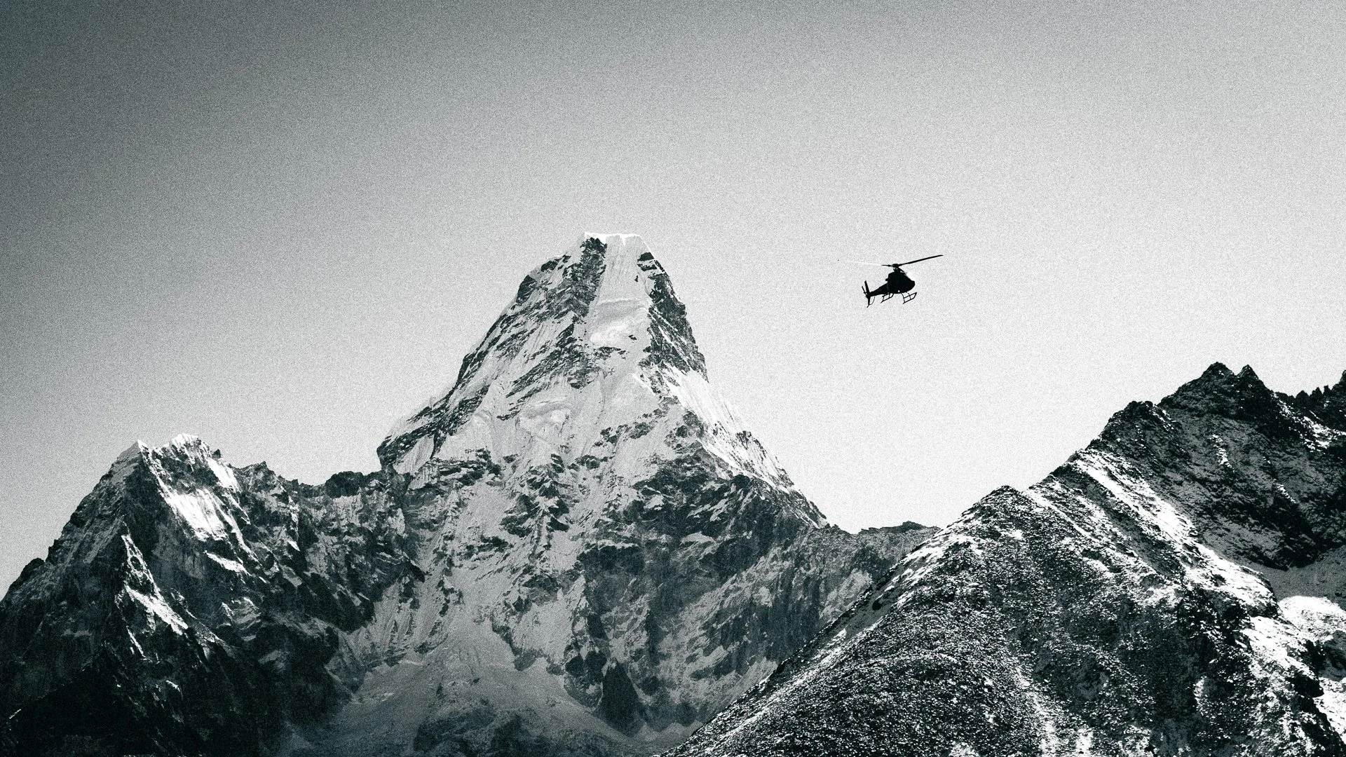 A black and white photo of a snow-capped mountain and smaller mountains below. A helicopter is flying in the sky to the right of the mountain peak.