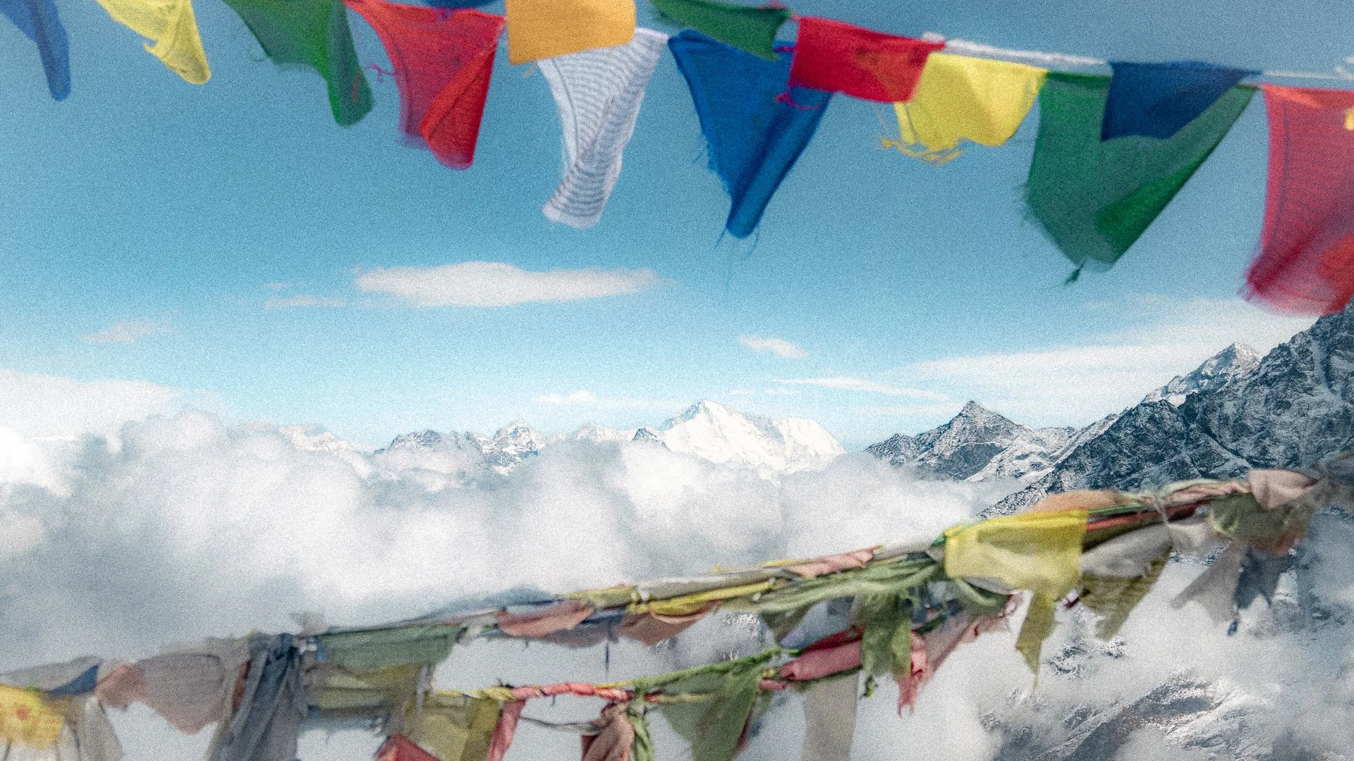 Colorful prayer flags hanging in the foreground with snow-capped mountains and a clear blue sky in the background.