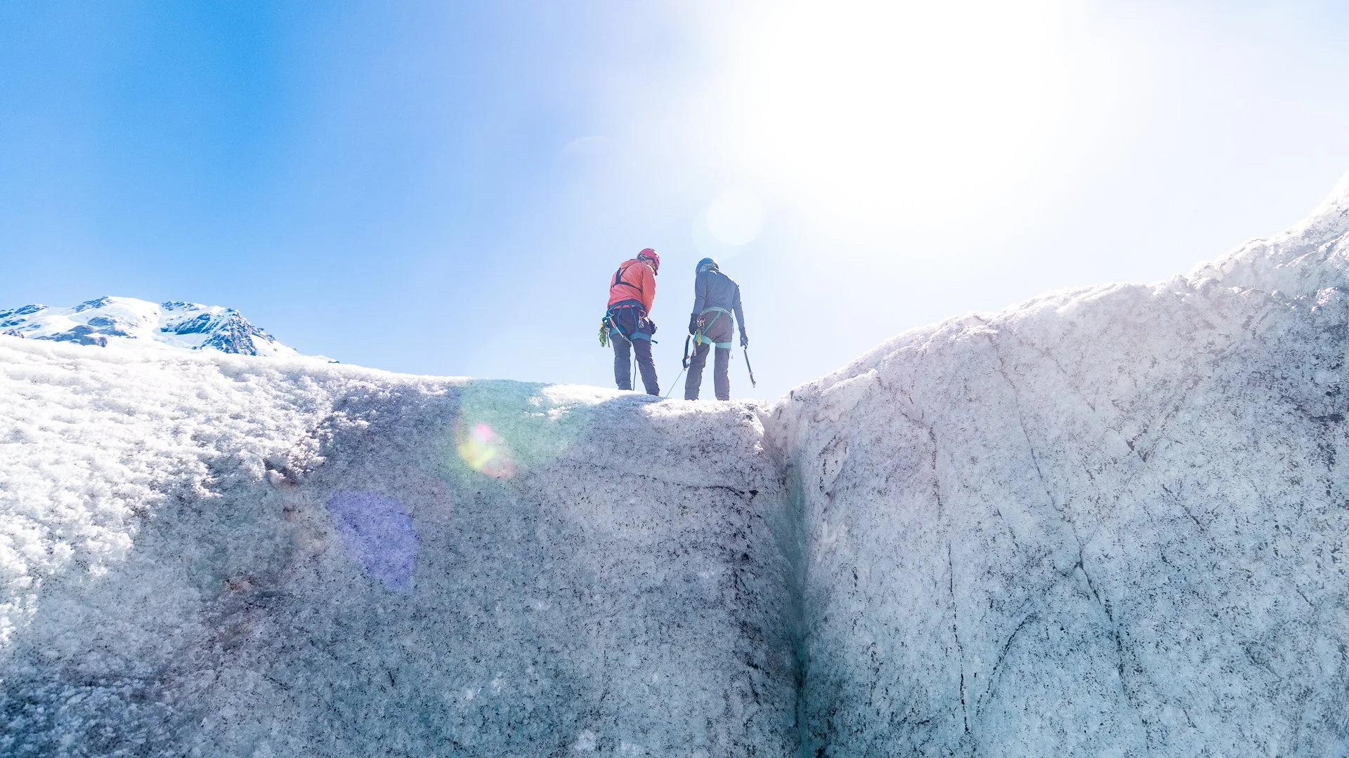 Two climbers standing on top of a glacier, looking at a mountain in the distance, under a bright sky.