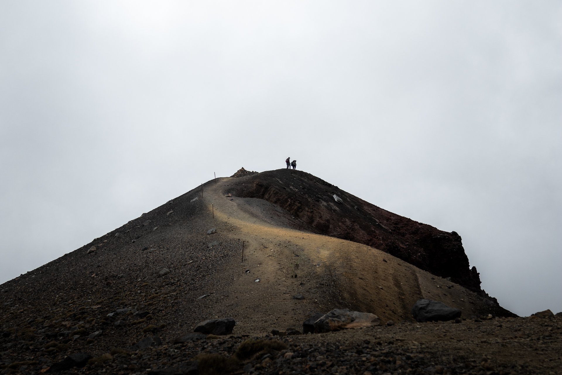 Two hikers standing on a mountain ridge with a dirt trail, dark rocky terrain, and sparse vegetation, under a cloudy sky.