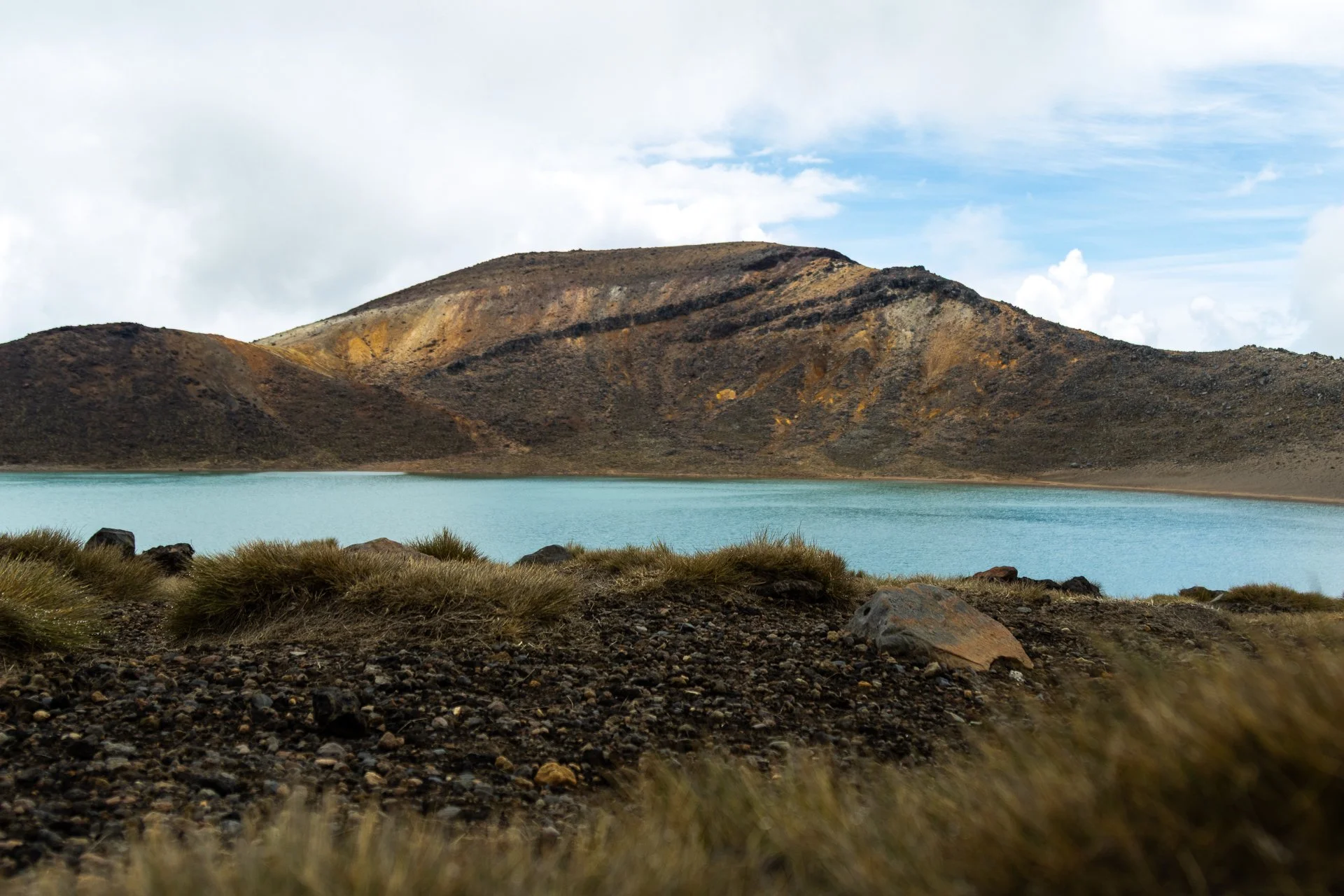 A mountain with a brown and rocky surface, partially cloudy sky, and a body of water in the foreground surrounded by grass and rocks.