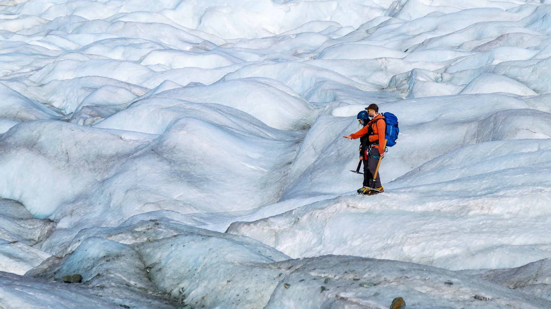 Two people, dressed in winter gear, standing on snow and ice on a glacier, with one pointing at something.