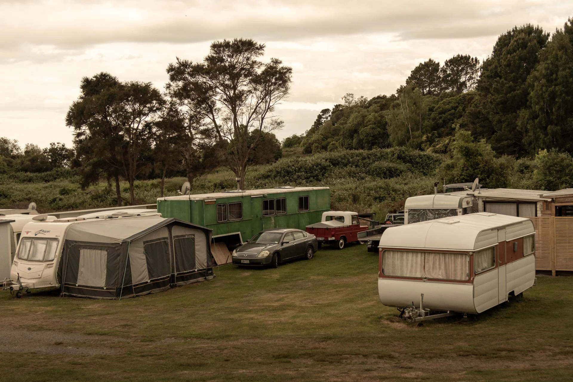 A campsite with multiple caravans, trailers, and cars parked on a grassy area, surrounded by trees and hills under a cloudy sky.