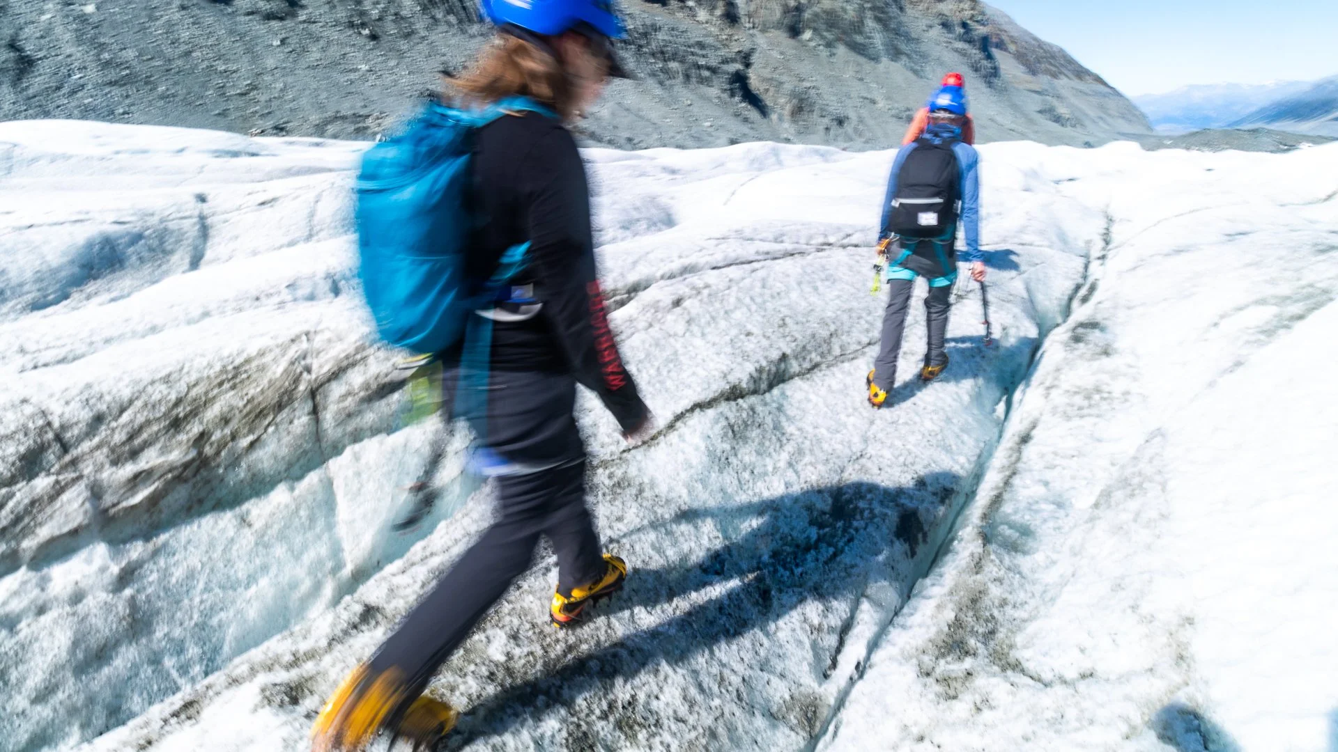 Two hikers with backpacks and helmets walking on ice glacier in mountainous landscape.