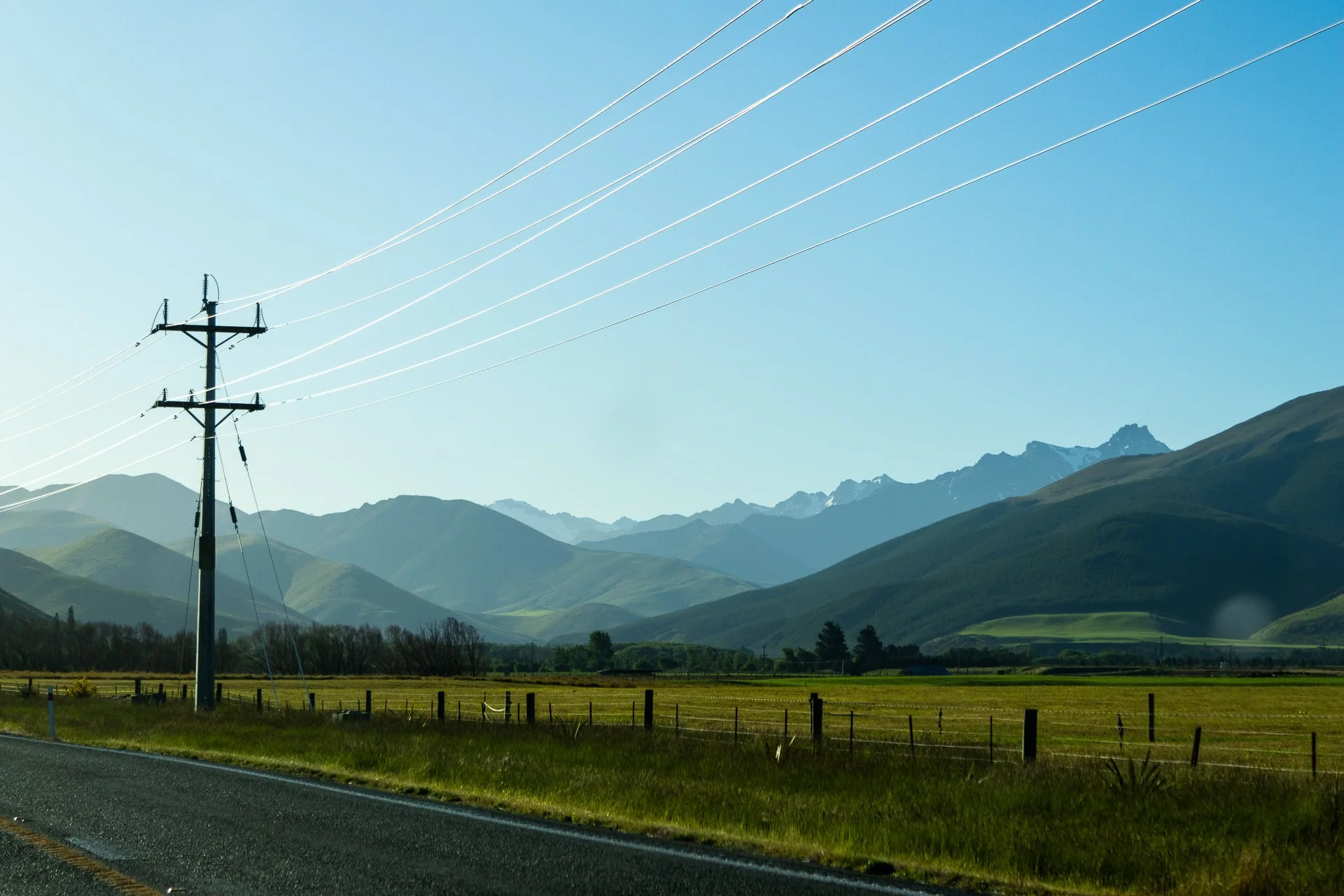 A landscape of rolling green hills with mountains in the background and clear blue sky. A utility pole with power lines runs diagonally across the scene, and a fence lines the roadside in the foreground.