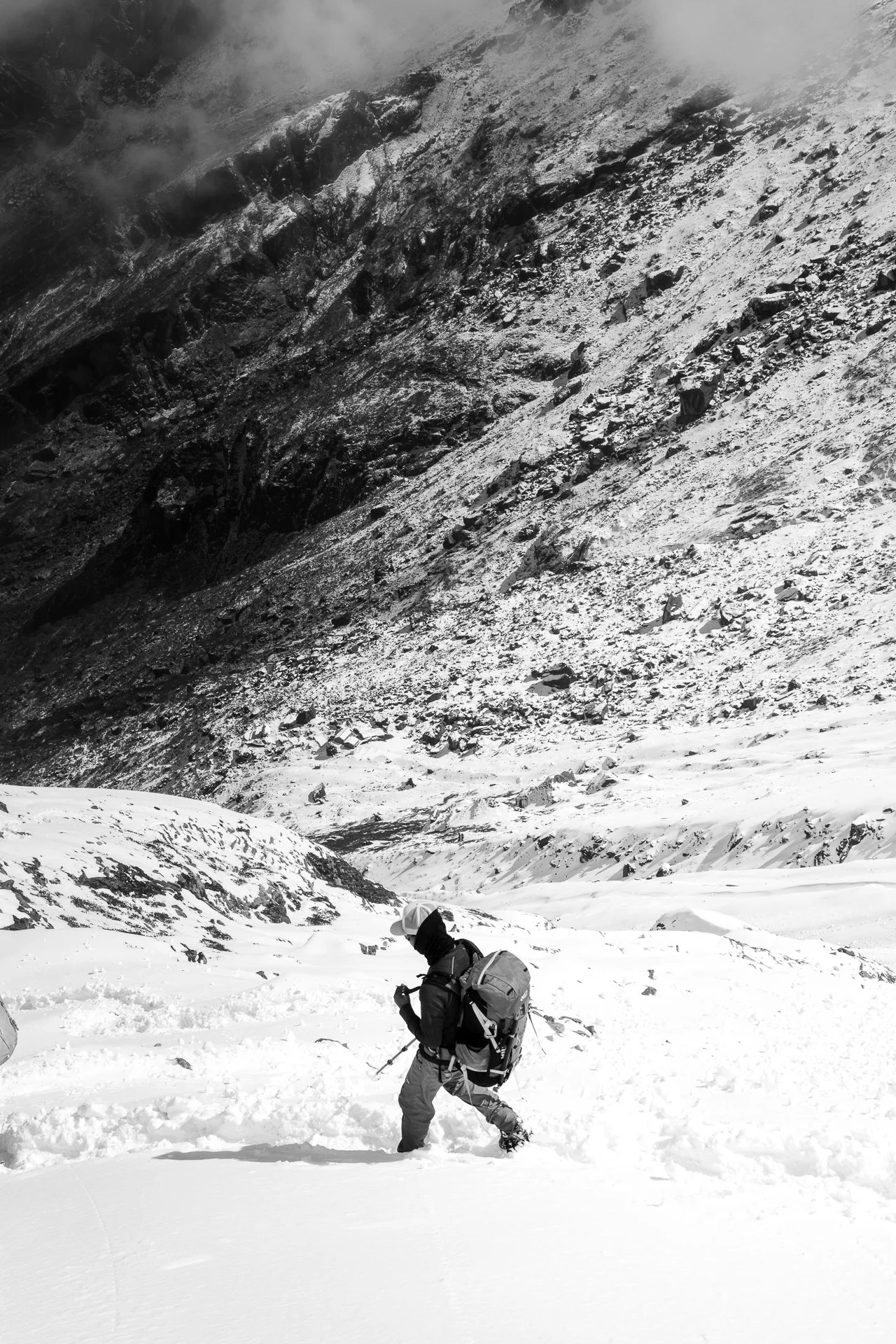 A person dressed in winter gear, carrying a backpack and hiking poles, ascending a snow-covered mountain slope surrounded by rugged rocky terrain.