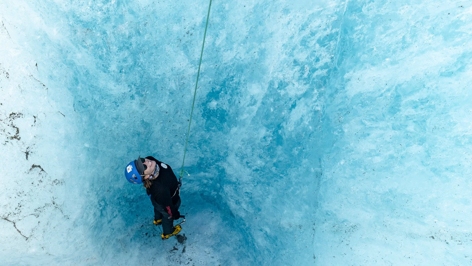 A person standing on an icy or snowy surface, wearing a blue helmet, black jacket, and yellow boots, looking up while holding an ice axe, with a safety rope attached to their harness.