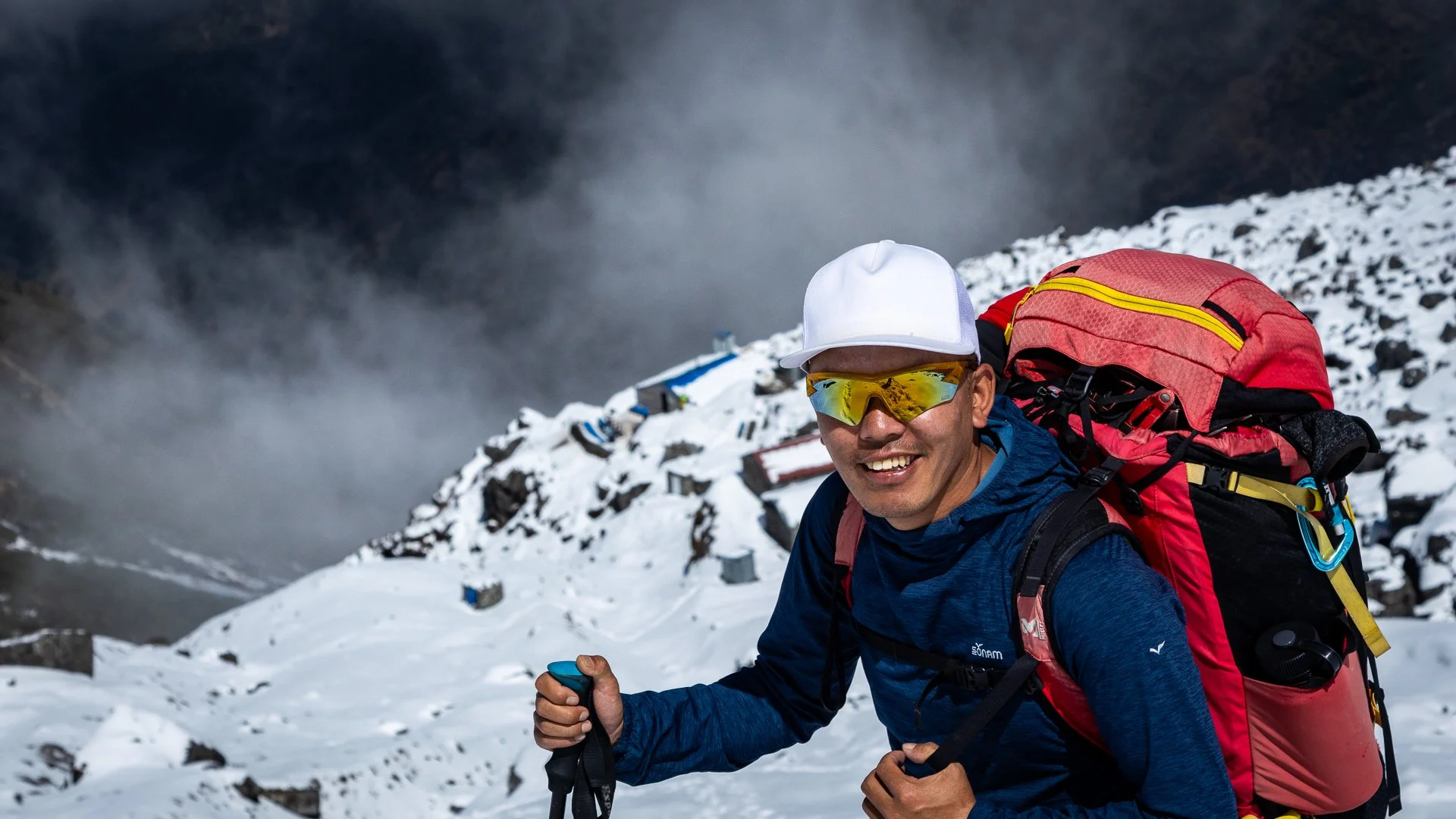Man with a red backpack and yellow sunglasses hiking on snowy mountains.