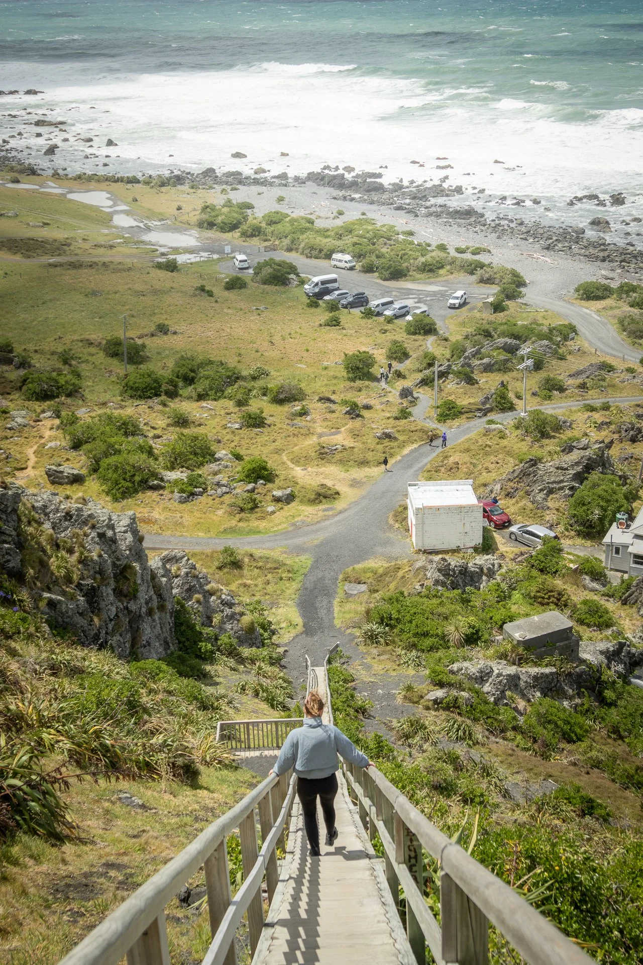 A person walking down a wooden staircase on a hillside towards a scenic view of a rocky beach, parking lot with cars, and the ocean in the background.