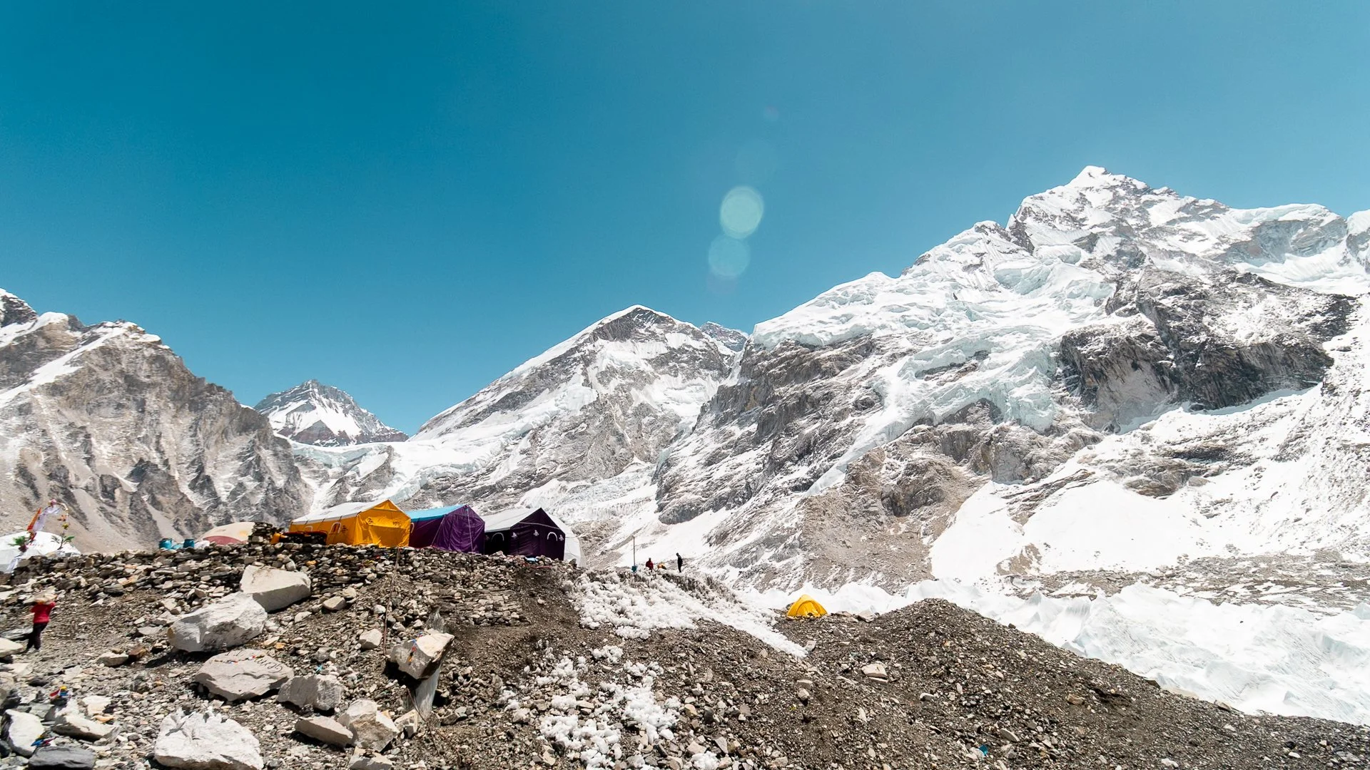 Mountain campsite with colorful tents set up on rocky terrain at base of snow-capped peaks under clear blue sky.