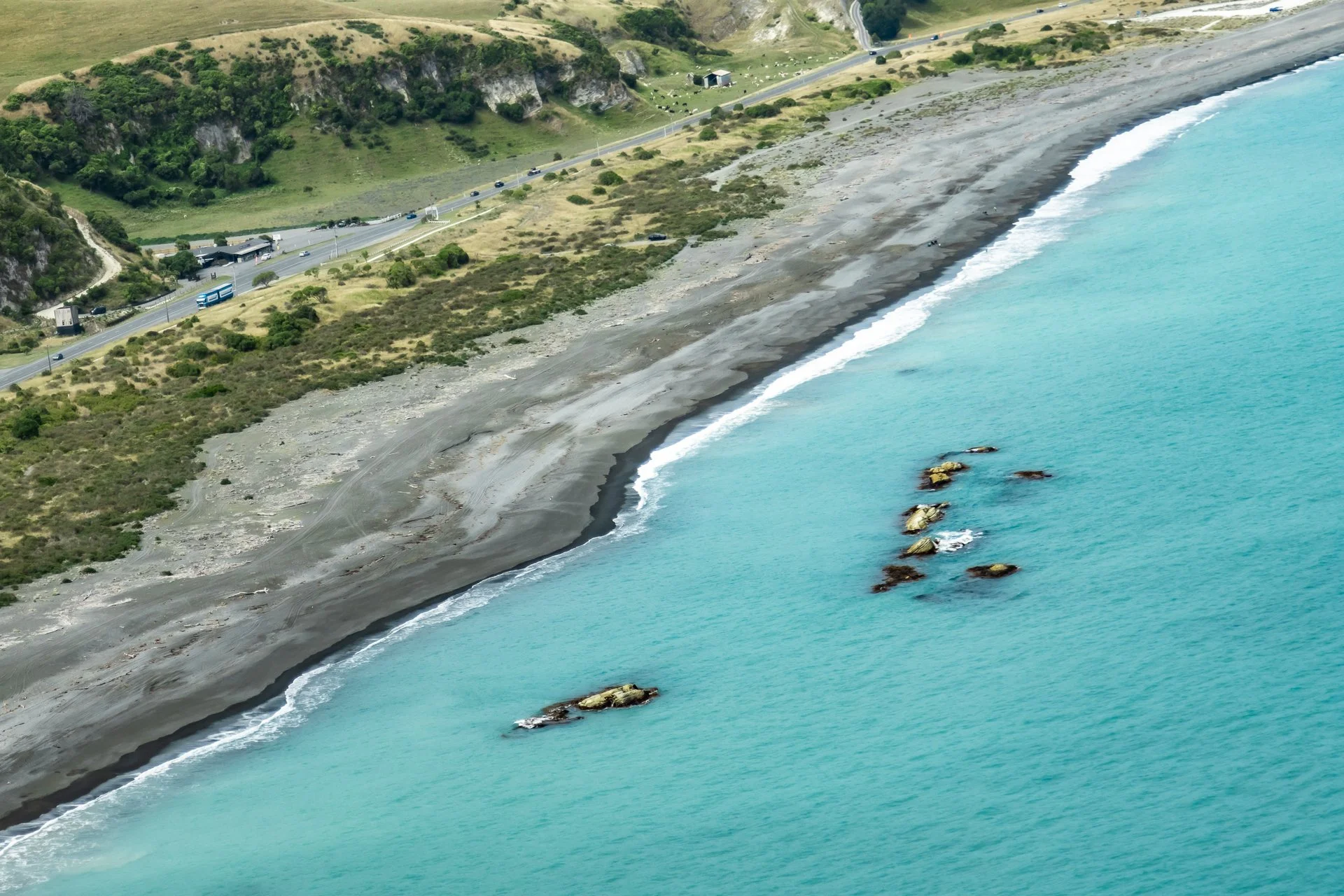 Aerial view of a coastal highway running along a rocky shoreline with clear blue ocean water and a few rocks in the sea.