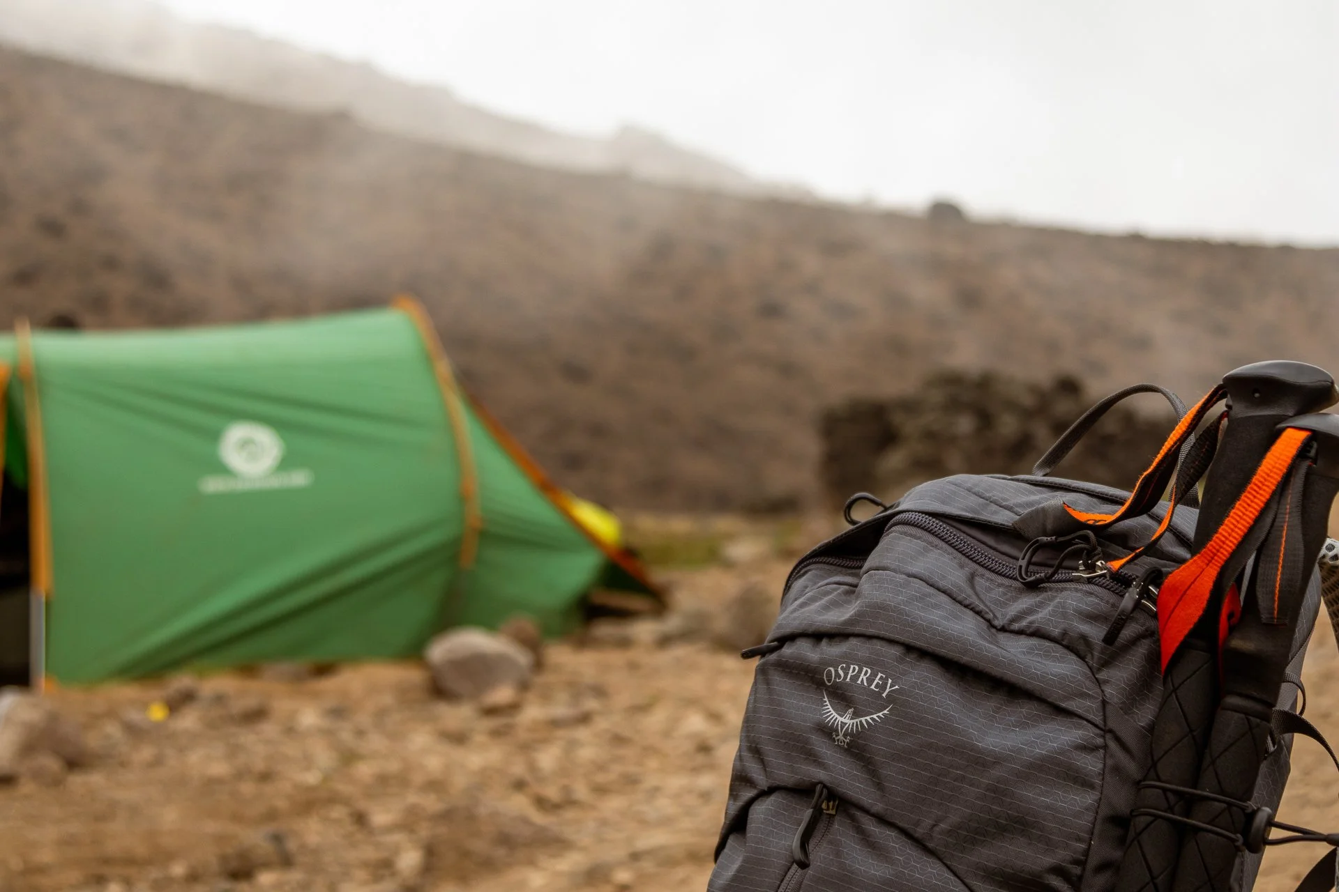 Camping gear, including a black backpack with orange straps and a green tent, set up on rocky ground in a mountainous outdoor setting with foggy weather.