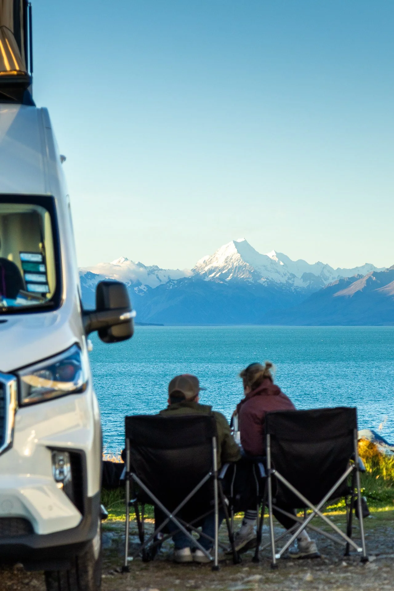 People sitting in camping chairs outdoors near a large body of water with mountains and snow-capped peaks in the background, and a partial view of a white vehicle.