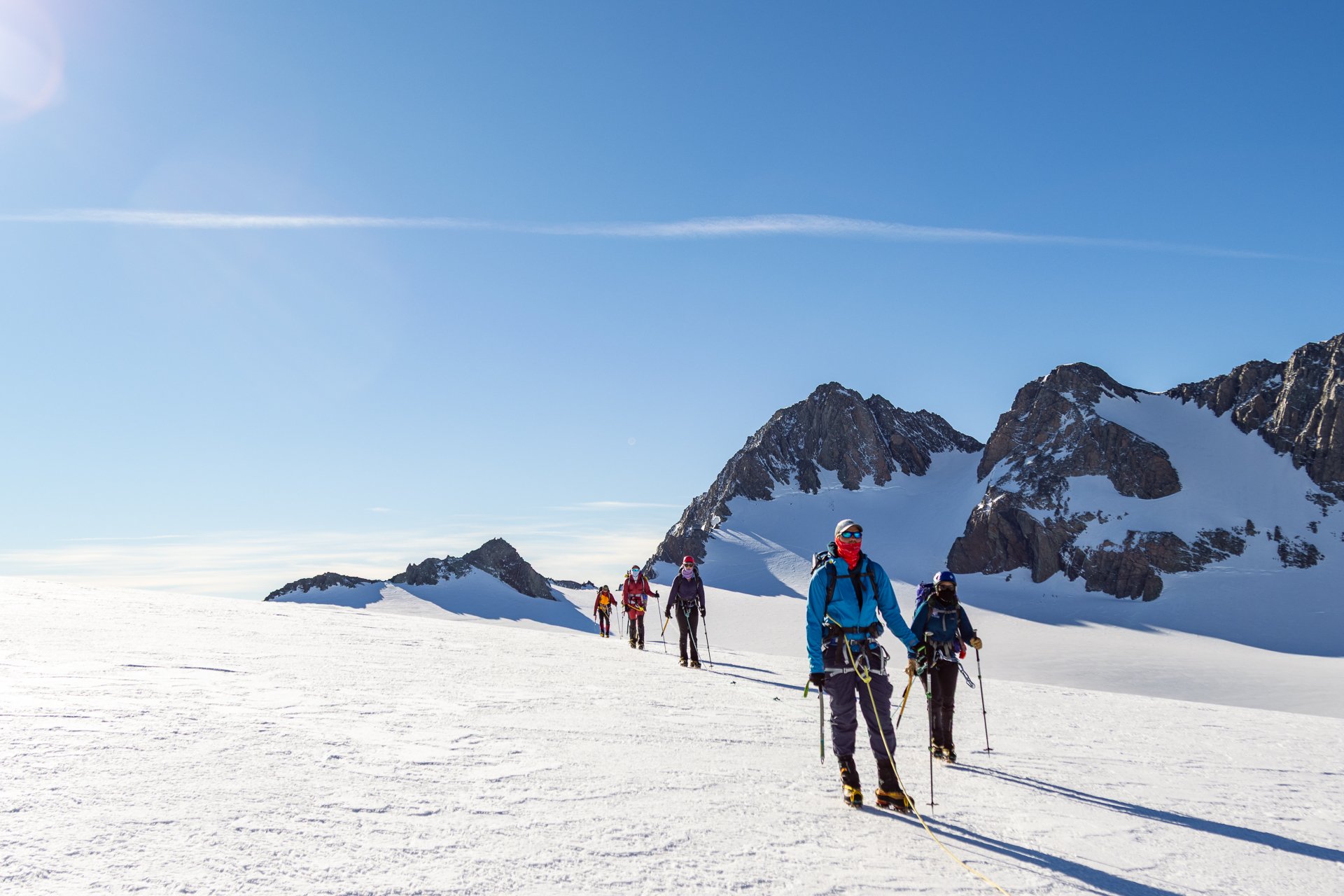Group of skiers ascending a snowy mountain with rugged peaks in the background under a clear blue sky.
