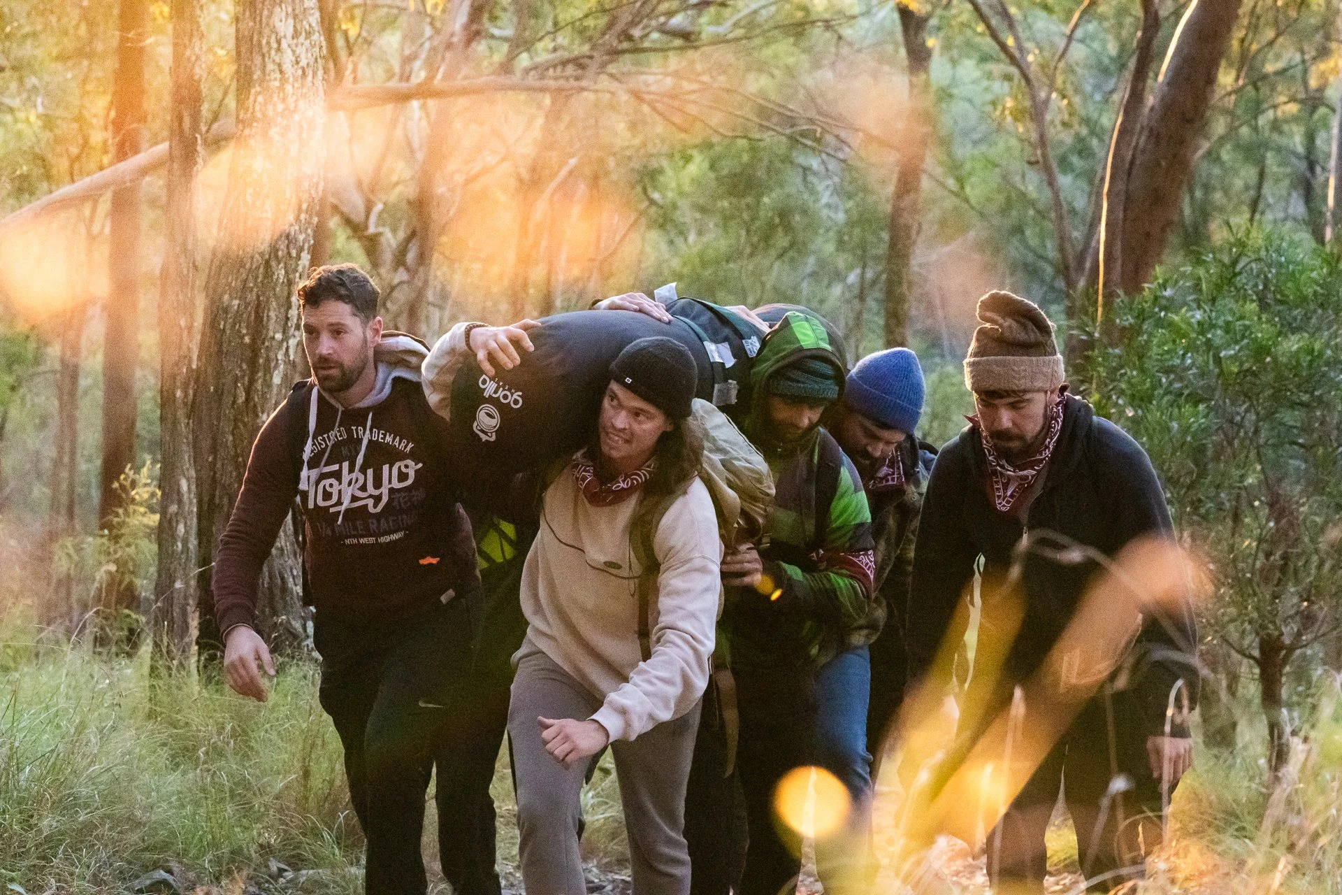 A group of five people hiking through a wooded forest trail during sunset. They are wearing outdoor clothing and carrying backpacks.