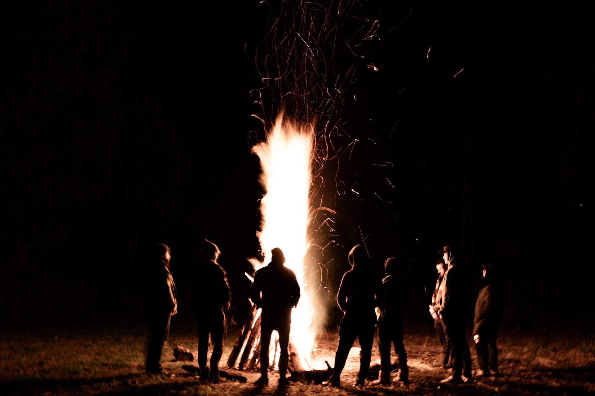 A group of people standing around a bonfire at night, watching sparks and flames rise into the dark sky.
