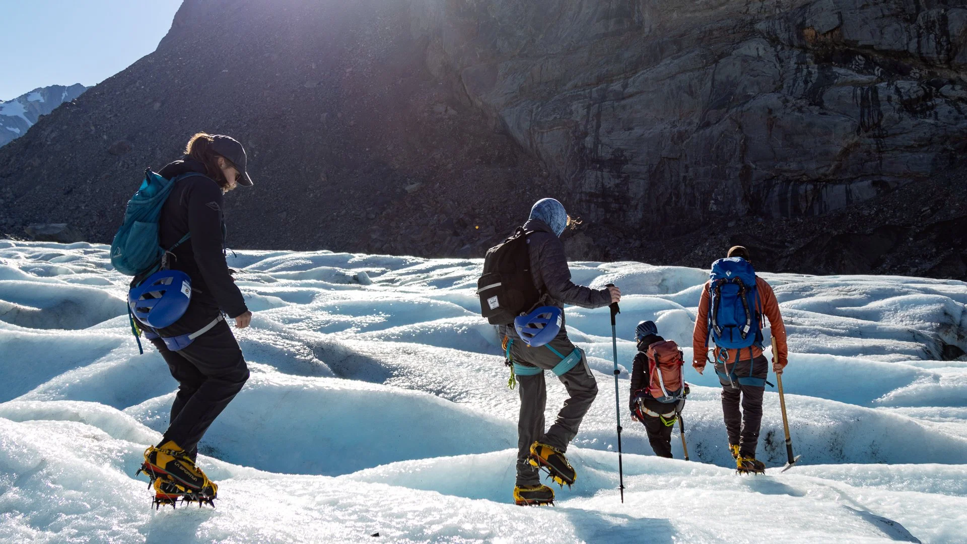 Four explorers wearing winter gear and ice crampons trek across a snow-covered icy terrain with backpacks, hiking poles, and helmets, surrounded by snow-capped mountains and a steep rocky cliff in the background on a clear day.