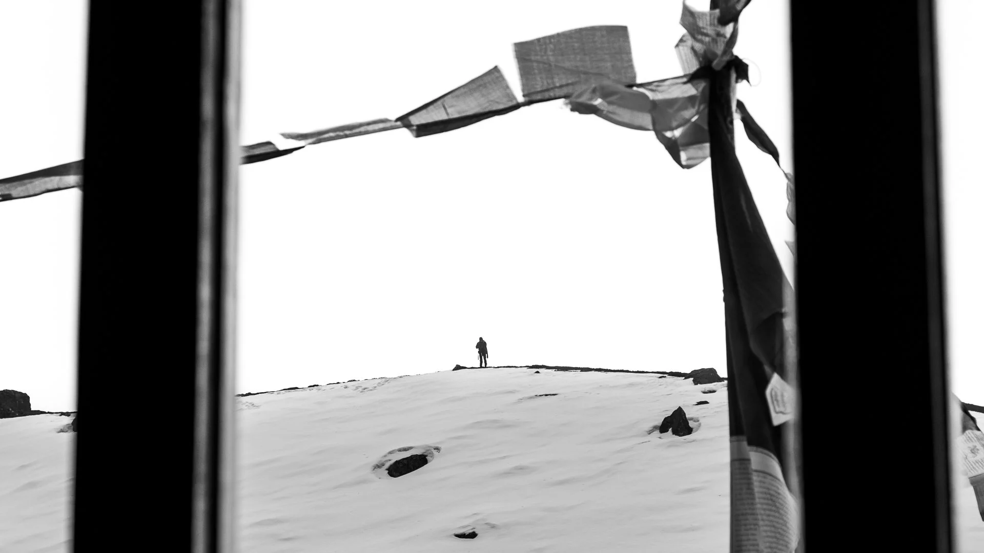 Black and white photo of a person standing on a snowy hill, taken from inside a structure with dark vertical frames and shreds of fabric hanging, possibly prayer flags, in the foreground.
