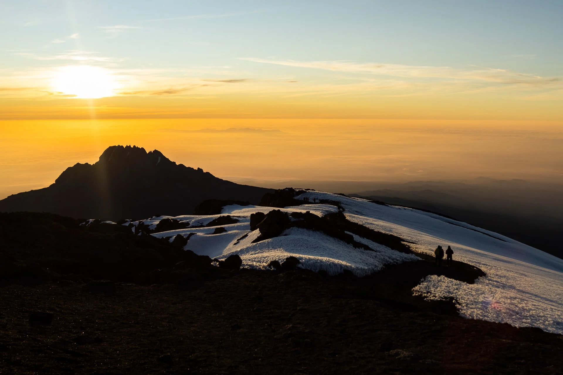 Sunset over a mountain landscape with snowy patches, silhouetted hikers on the trail, and a colorful sky with scattered clouds.