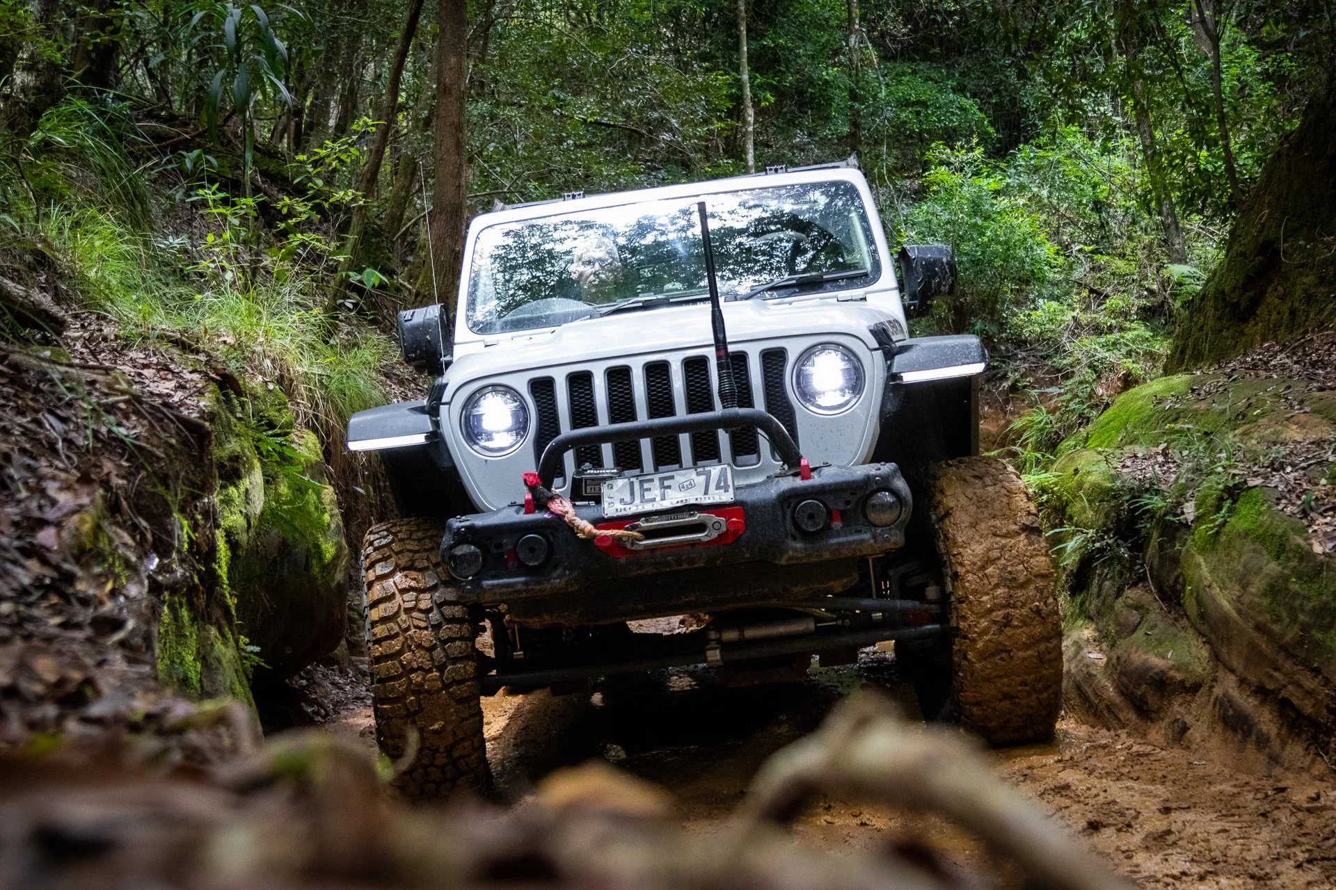 A white off-road vehicle with large tires navigating through a narrow, muddy trail surrounded by dense green forest.