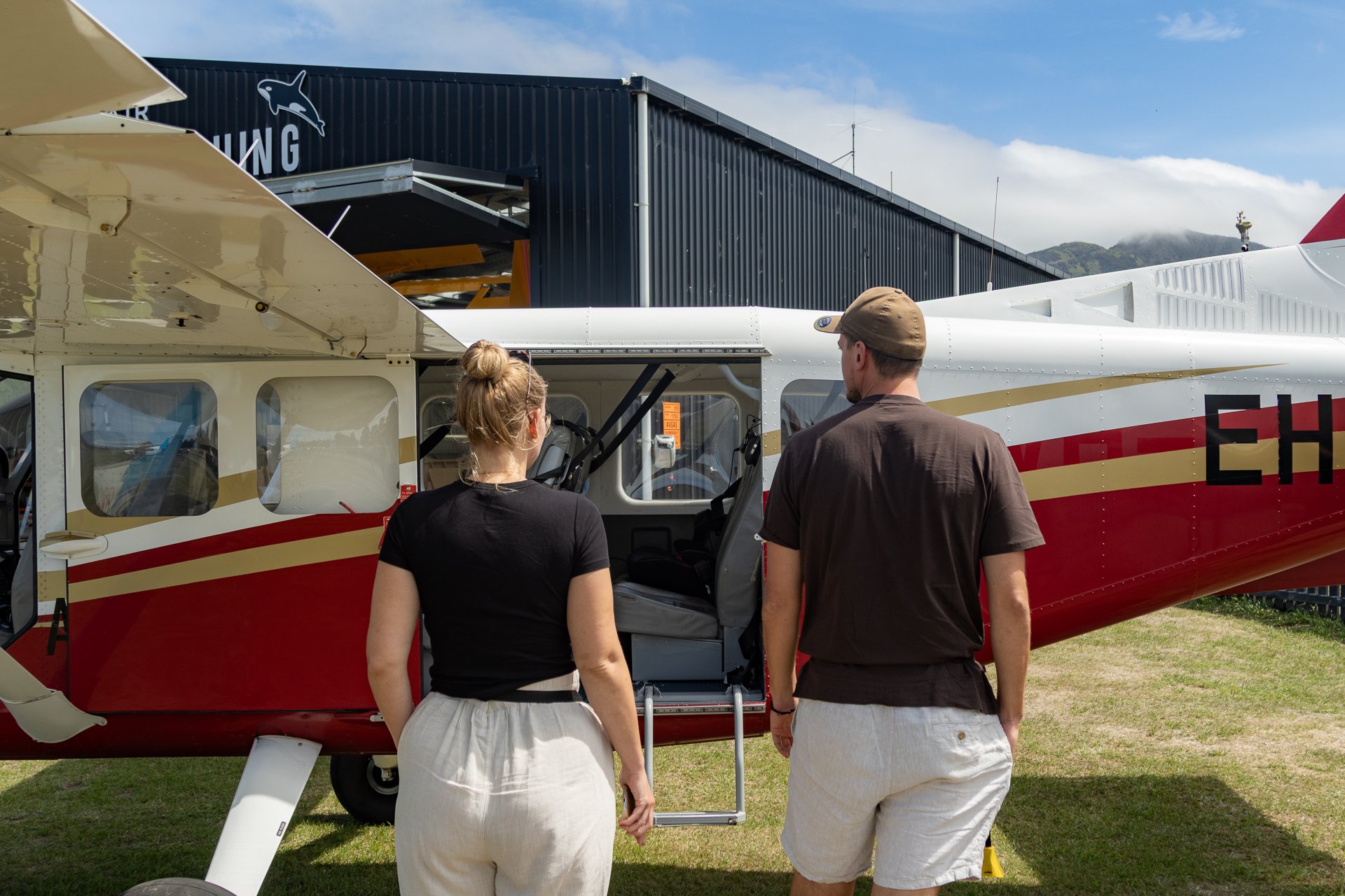 Two people, a woman with blonde hair in a bun wearing a black top and white skirt, and a man with a brown cap and shirt with white shorts, are standing next to a small red and white aircraft on an airfield. The aircraft has an open door with stairs l