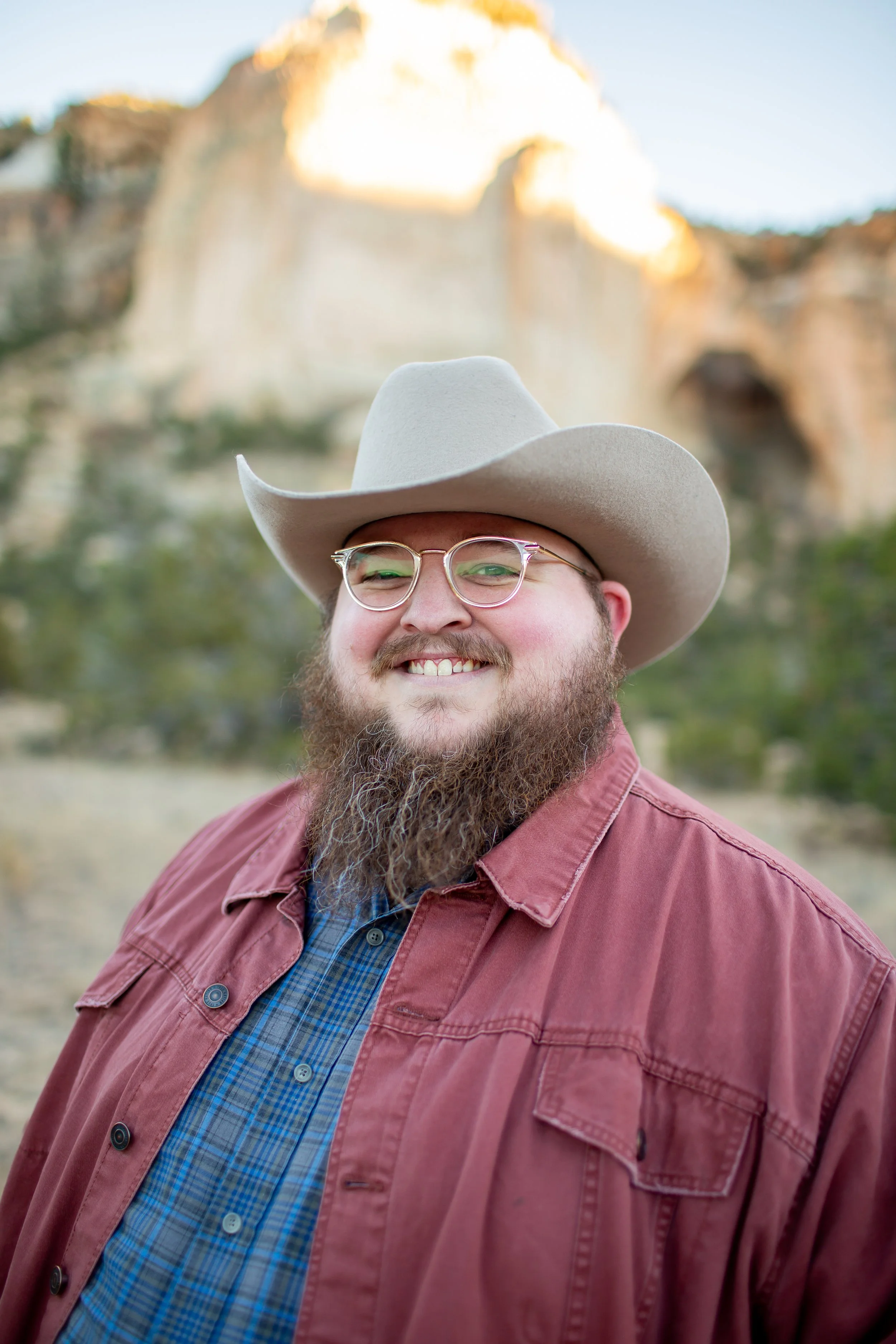 Man(Giles) with a beard, glasses, wearing a white cowboy hat, plaid shirt, and red jacket smiling outdoors with rocky cliffs and trees in the background.