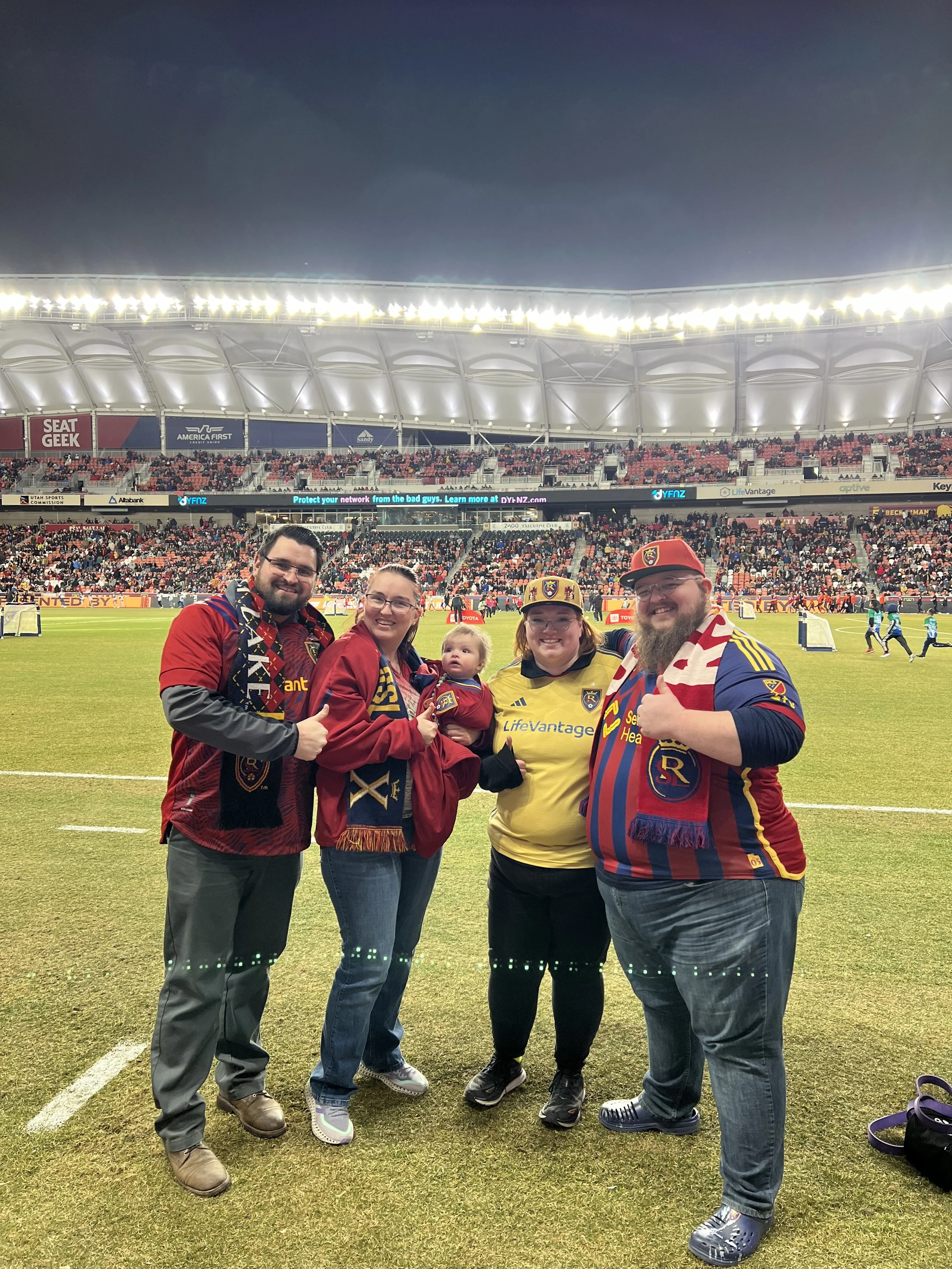 Giles, Kayla, and friends on the field at night during a Real Salt Lake game after being featured at halftime, wearing soccer jerseys and scarves, with a stadium full of spectators in the background.