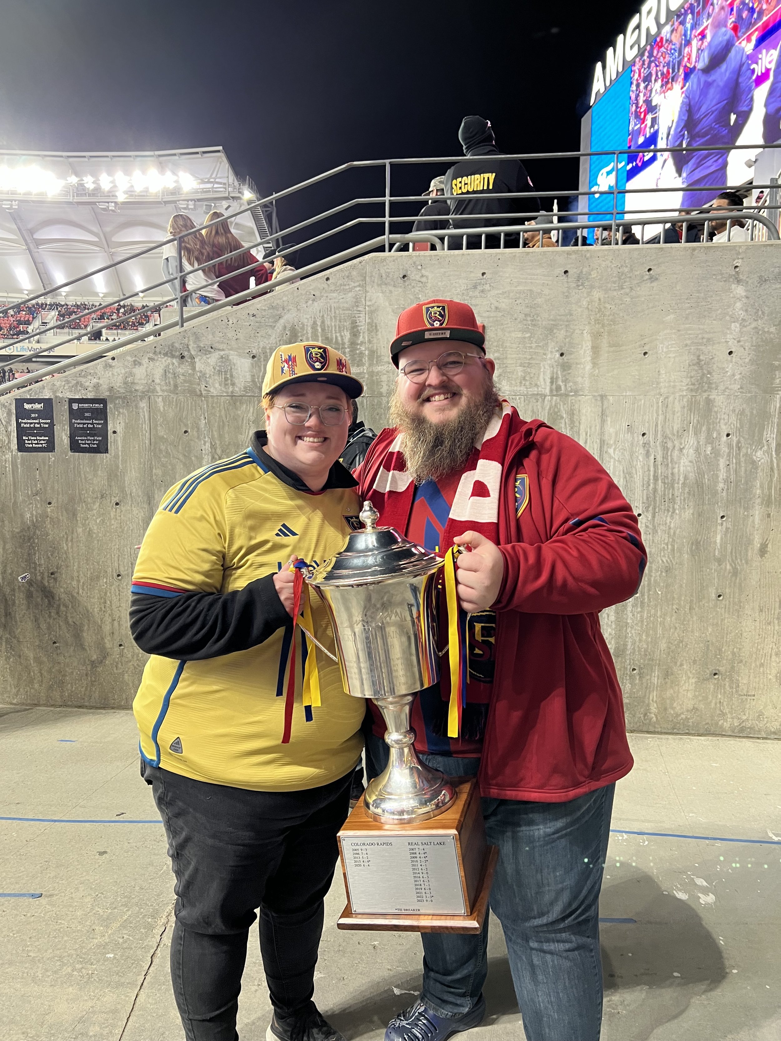 Giles and Kayla holding the Rocky Mountain Cup at America First Field during halftime activities. They are smiling, standing in front of a concrete barrier with a stadium and large screen in the background.
