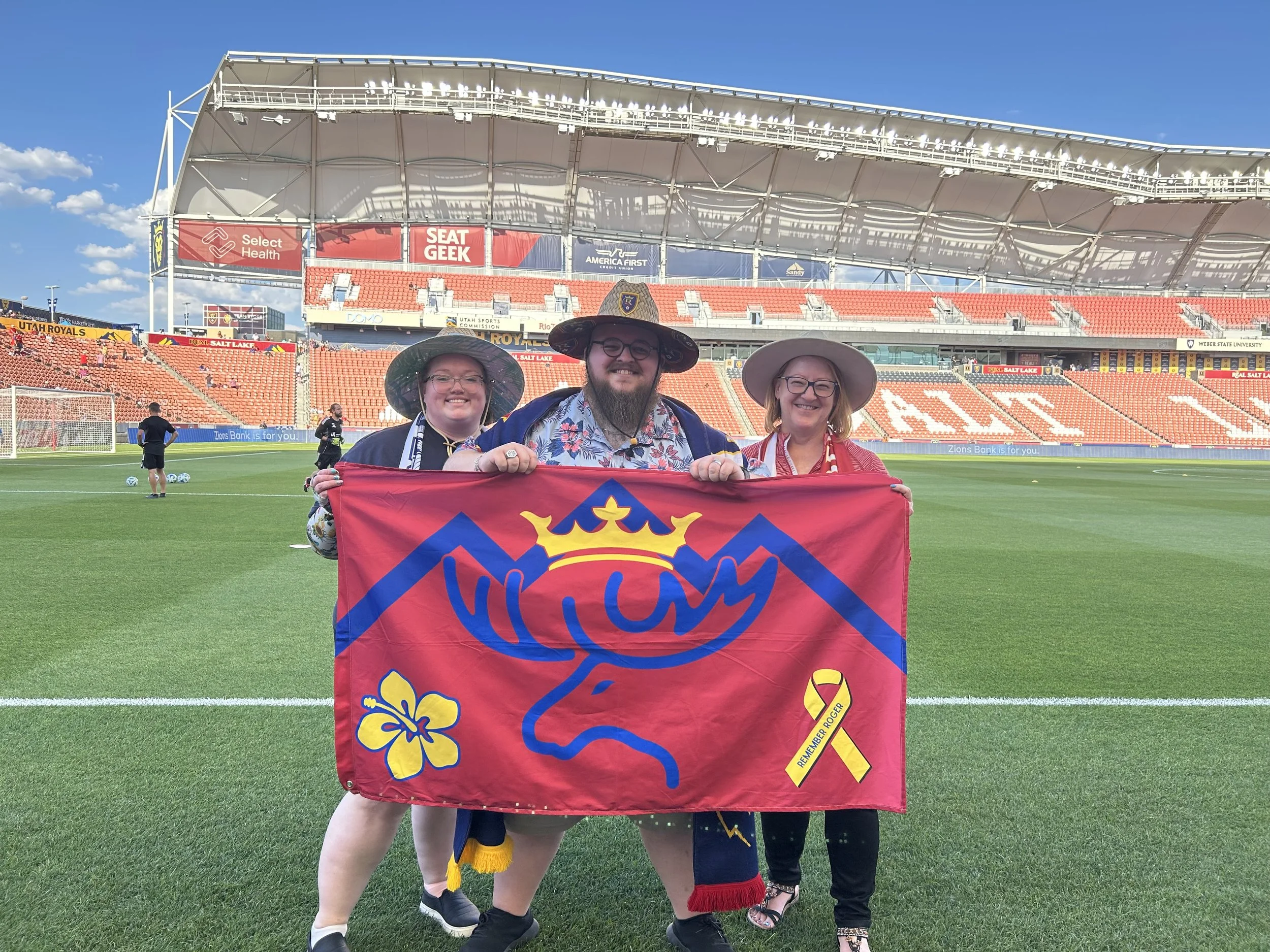 Giles, Kayla, and Linda standing on the field at America First Field, holding a flag with a logo and a yellow ribbon that reads 'Remember ROGER'. They are smiling and appear happy.