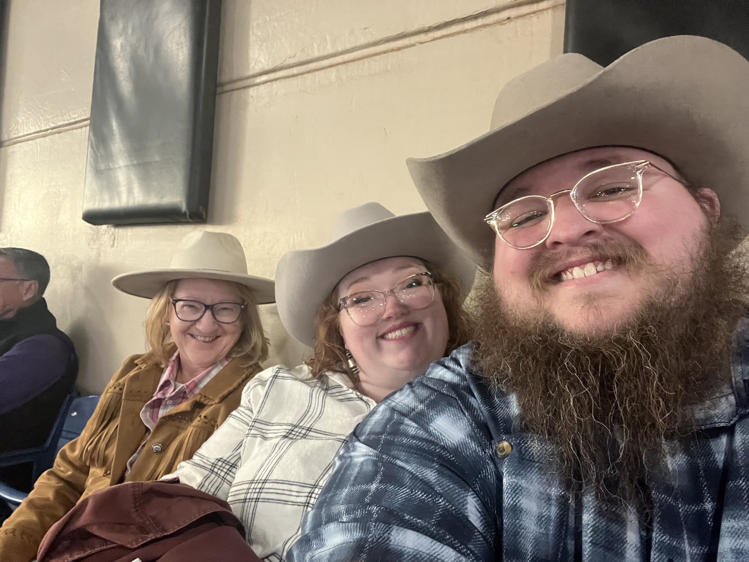 Giles, Kayla, and Giles' mom Linda smiling at the camera at the PRCA Wilderness Circuit Finals. All wearing large cowboy hats and glasses.