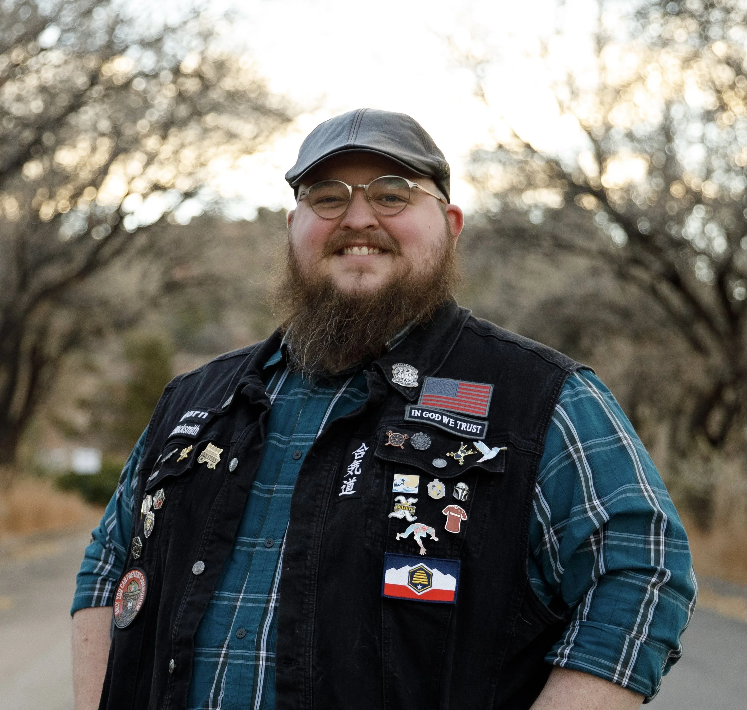 Giles with beard and glasses wearing a cap and a black vest with numerous patches, pins, and badges, standing outdoors during sunset with trees in the background.