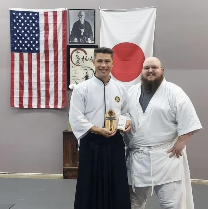 Giles presenting a hand forged incense burner to a visiting sensei at  an Aikido dojo with American and Japanese flags on the wall behind them.