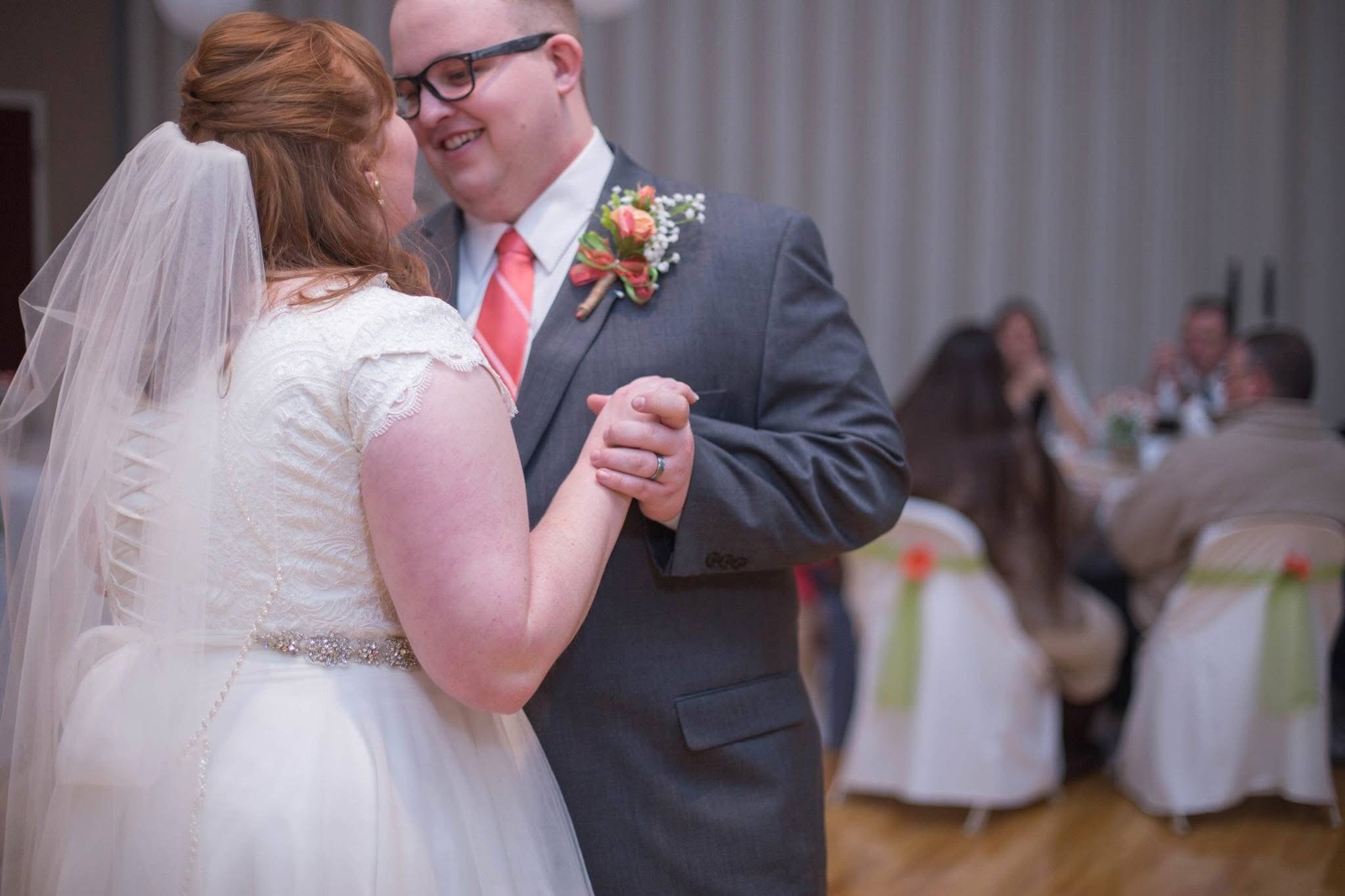 Giles and Kayla are dancing at their wedding reception, holding hands and smiling at each other, with guests seated at tables in the background.