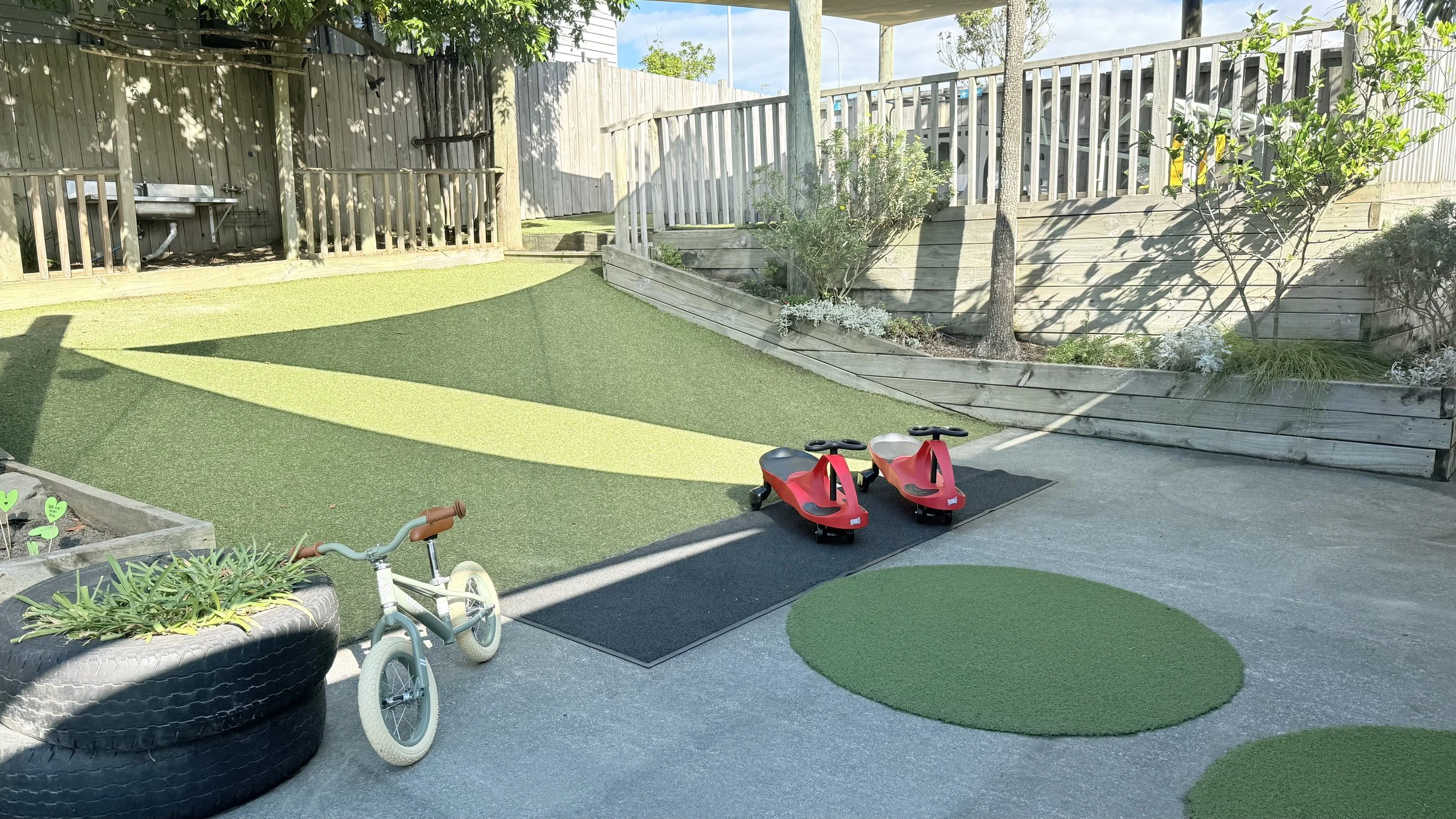 Child's backyard play area with artificial grass, a small bicycle, and two red ride-on toy cars on a rubber mat. There is a tire planter with plants, a small garden bed, and a wooden retaining wall with plants on top. A wooden fence surrounds the yard.