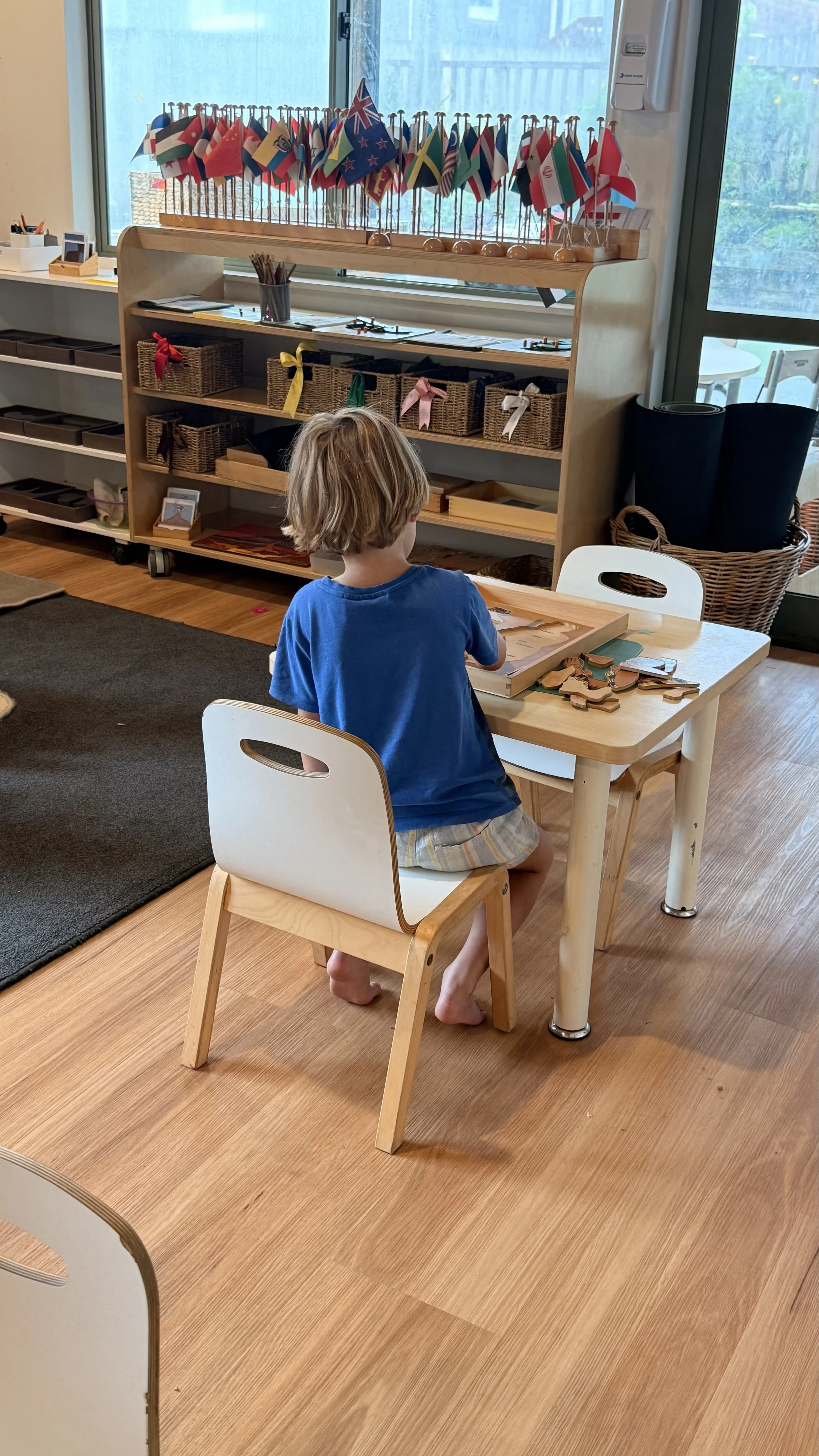 A young child with blond hair, wearing a blue shirt and shorts, sitting barefoot at a small wooden table playing with wooden puzzles in a classroom with shelves of baskets and children’s flags on display at Ponderosa Kids Albany