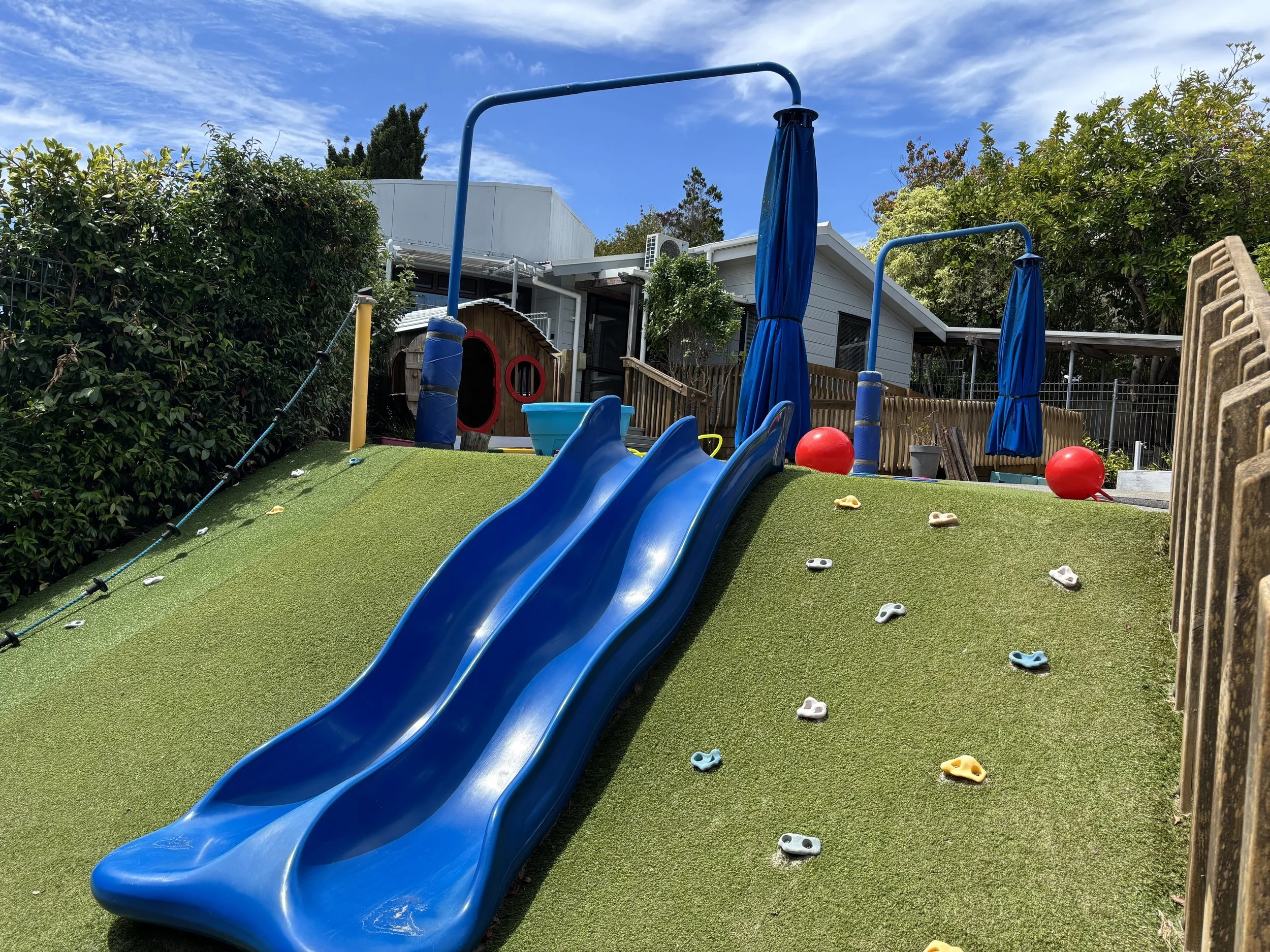 Children's playground with a blue slide, climbing holds on the grass, climbing poles with blue curtains, and a small playhouse in the background, surrounded by trees and a fence on a sunny day at Killarney Kids Bespoke Early Learning in Takapuna