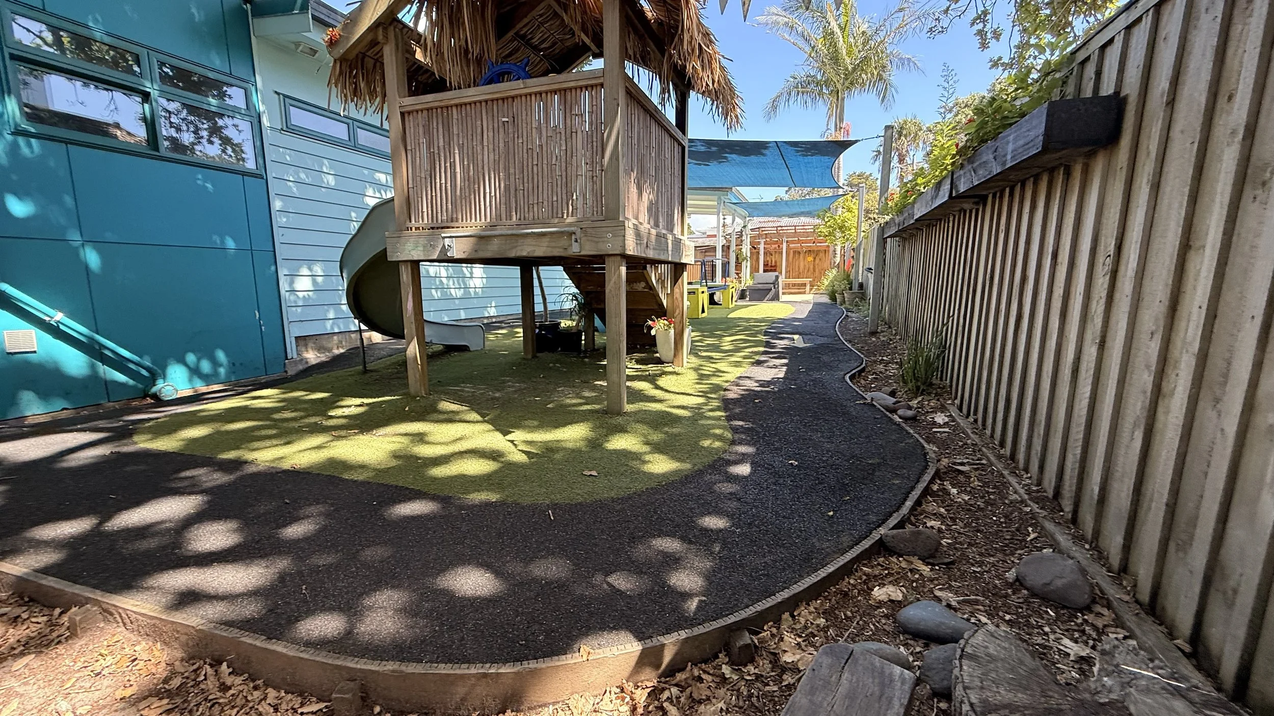 Backyard with a wooden playhouse, slide, and climbing structure, a curved black rubber pathway at Kiwi Kids Early Learning in Takapuna at Kids Group.