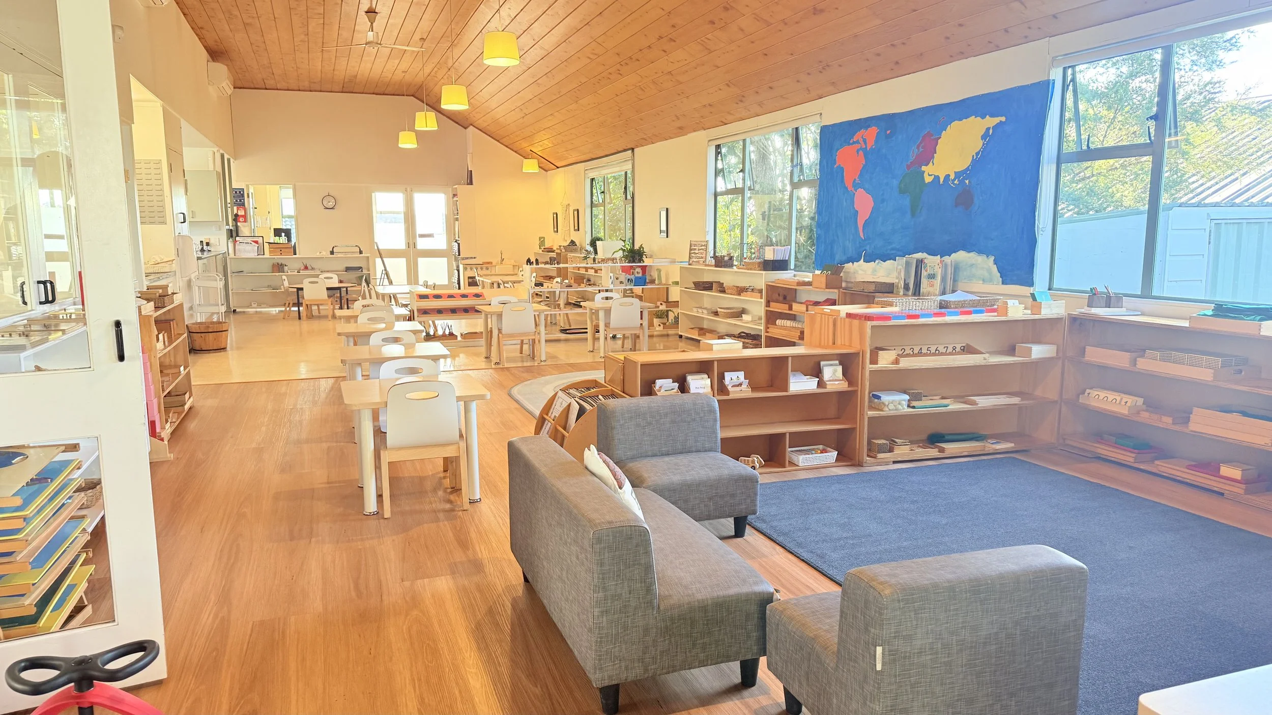 Bright preschool classroom with wooden floors, a wood-paneled ceiling, and large windows. Small tables and chairs are arranged throughout, along with shelves of educational materials and books at ponderosa kids Albany