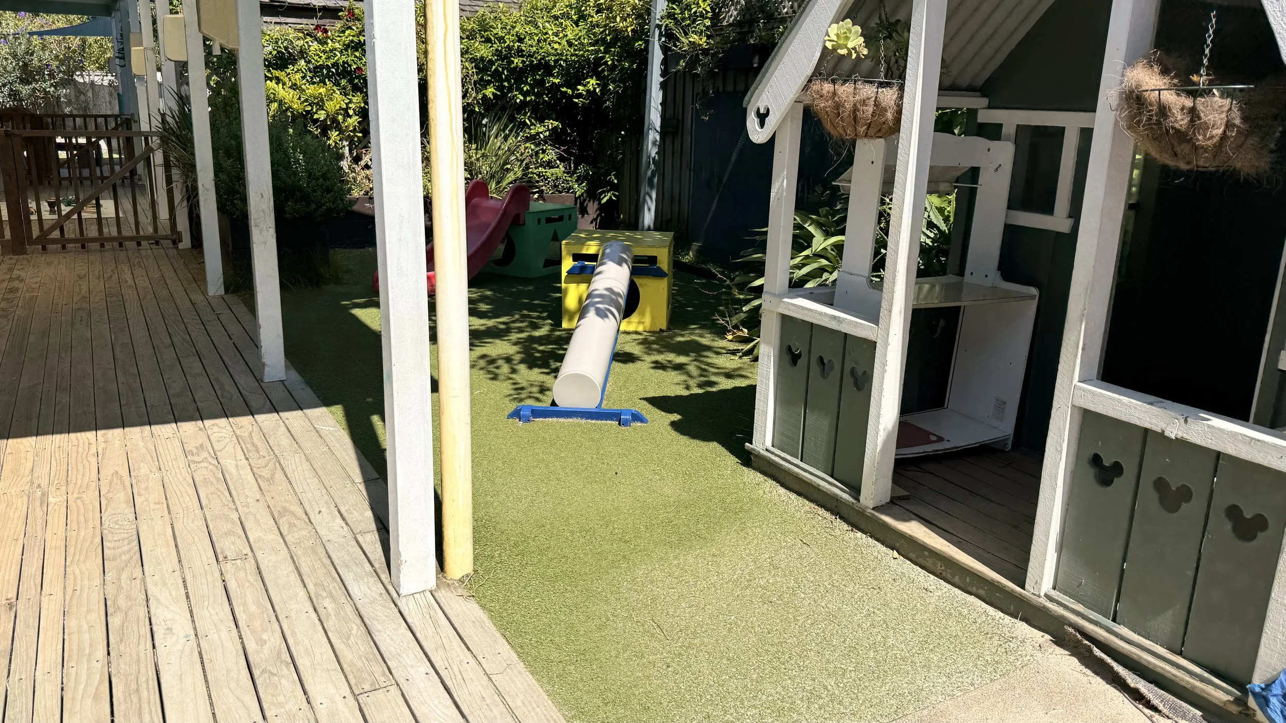 Children's outdoor play area with a slide, tunnel, and playhouse, surrounded by greenery and wooden fencing at Kiwi Kids Early Learning in Takapuna at Kids Group.