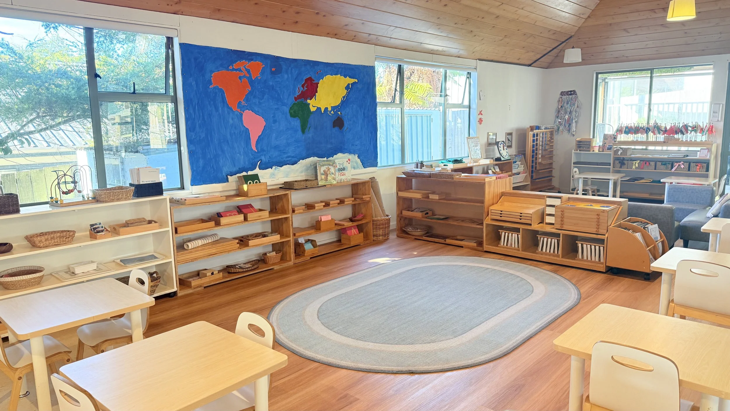 A tidy preschool classroom with wooden furniture, small tables and chairs, shelves with educational materials, a colorful world map mural on the wall, large windows letting in natural light, and a central oval rug on a wooden floor.