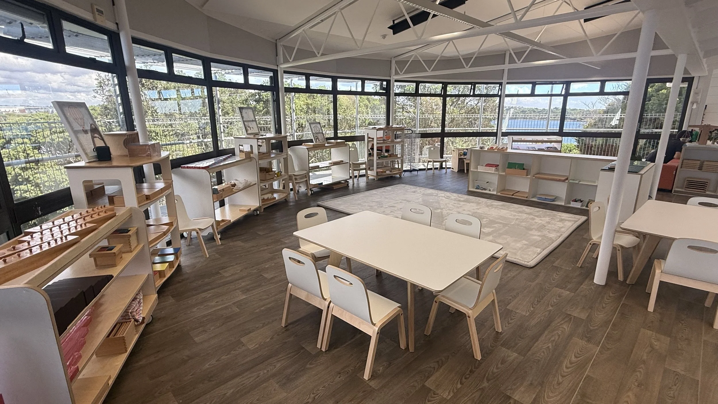 A bright classroom with large windows, white furniture, a central table with six chairs, shelves with educational materials, and a rug on the wooden floor.