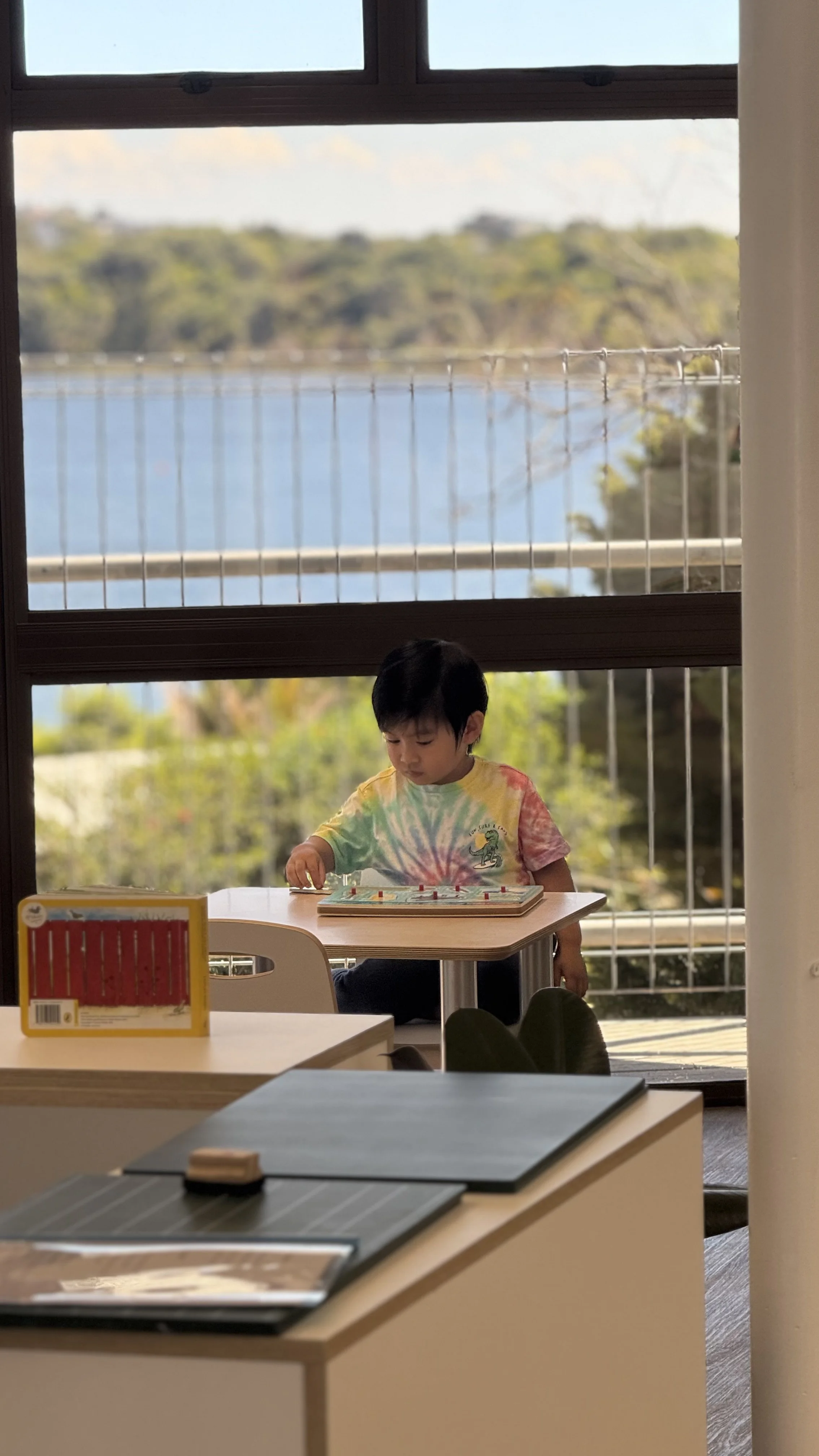 A young boy sitting at a small table playing a board game, with a large window behind him showing trees, water, and a fence outdoors at Killarney Kids Bespoke Early Learning in Takapuna at Kids Group.