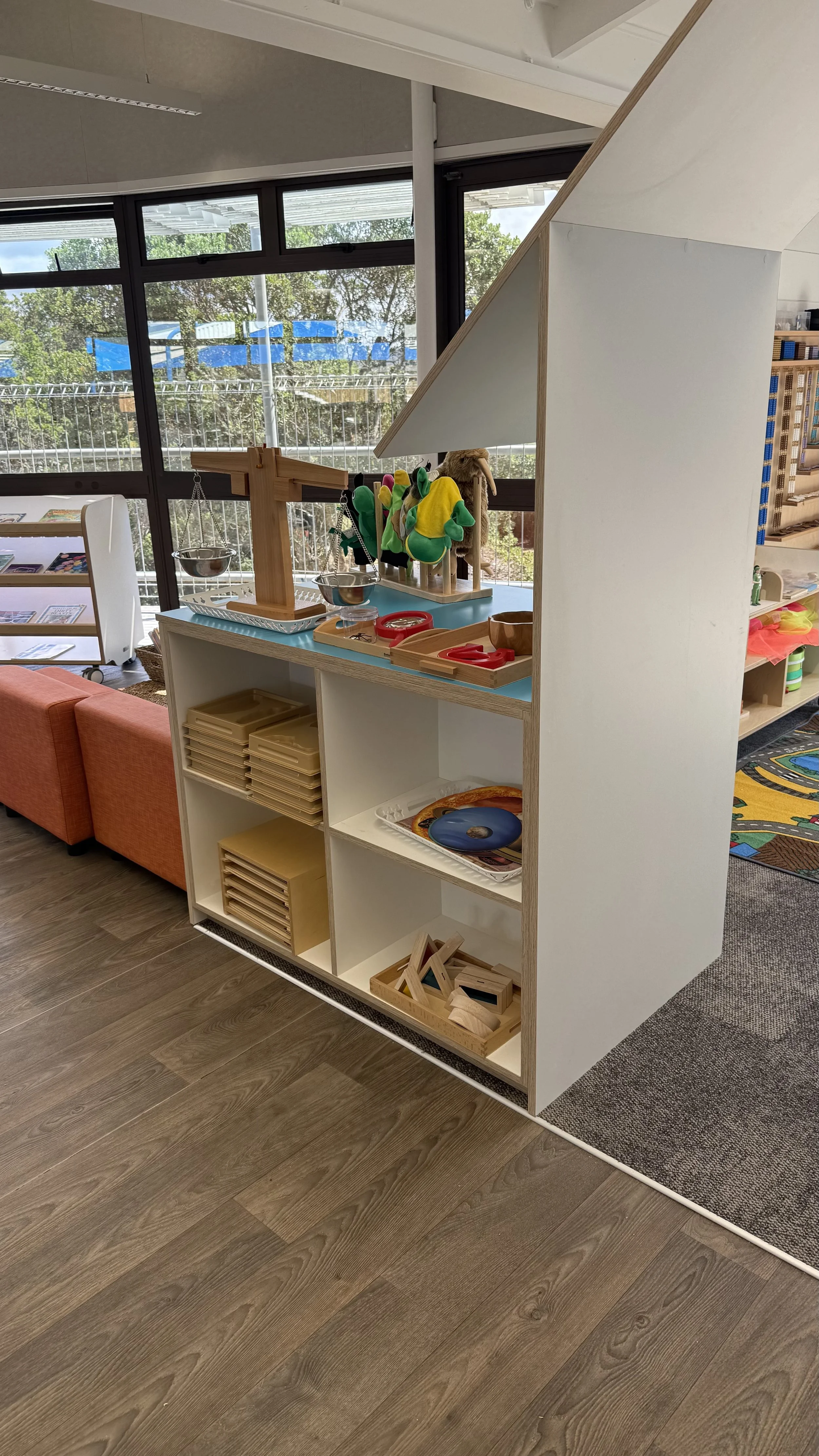 Inside main classroom, a white bookshelf with wooden trays and toys, including plush animals, wooden puzzles, and a tray with a doughnut-shaped object, near large windows with outdoor trees visible at Killarney Kids Bespoke Early Learning in Takapuna