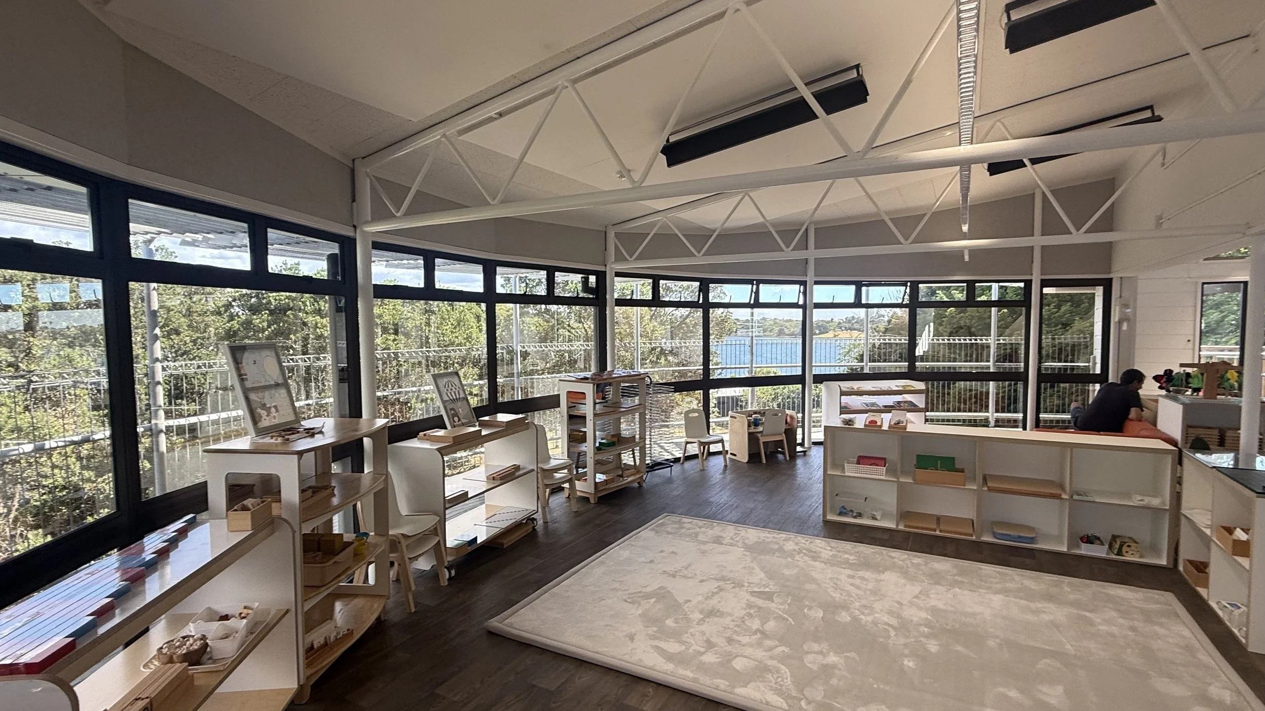 Interior view of a bright main classroom with large windows, white shelves, and a white rug, with a person seated working at a desk at Killarney Kids Bespoke Early Learning in Takapuna at Kids Group.